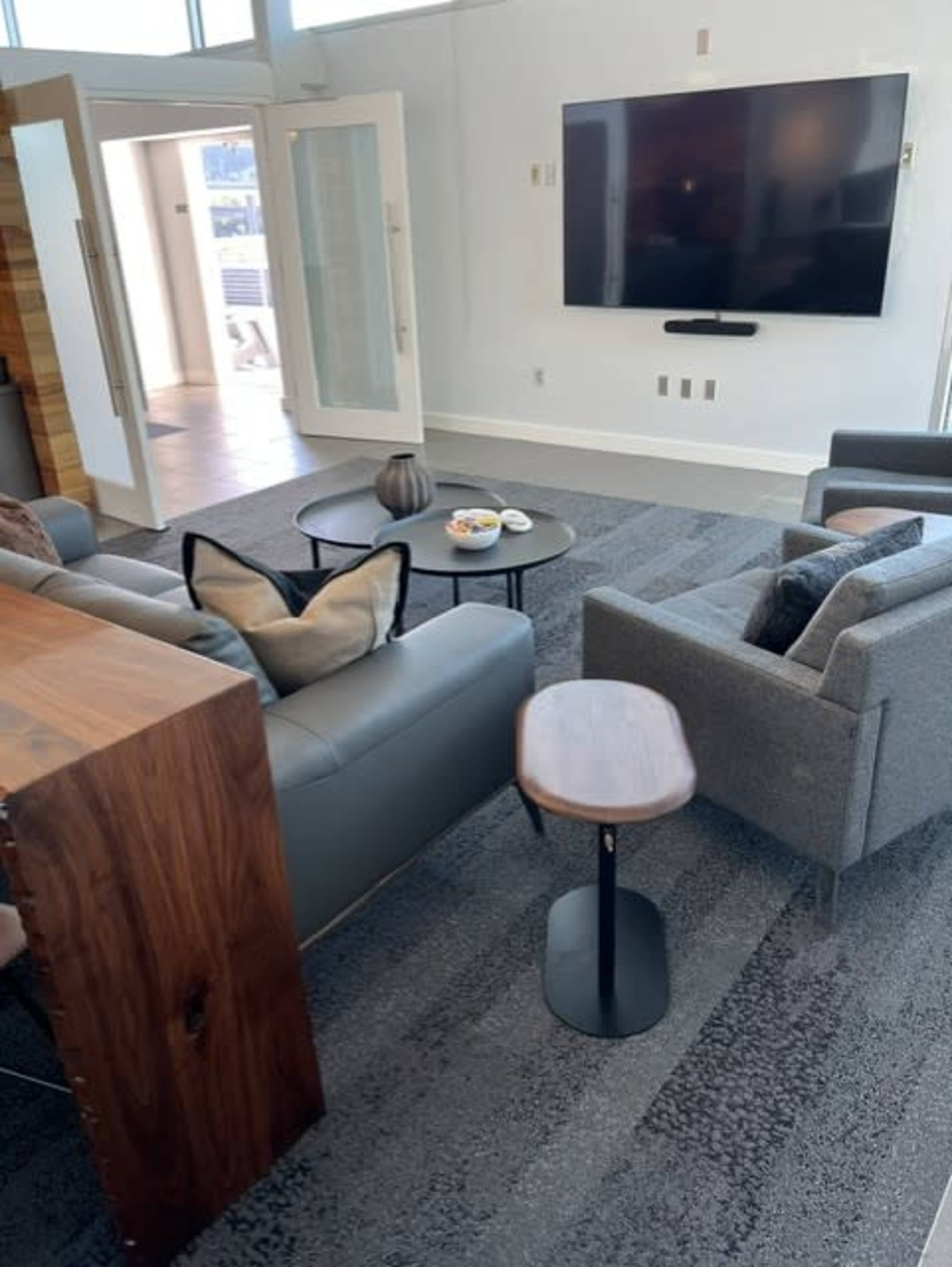 The image shows a modern living room with a gray sofa, two chairs, a coffee table, and a large television mounted on the wall.