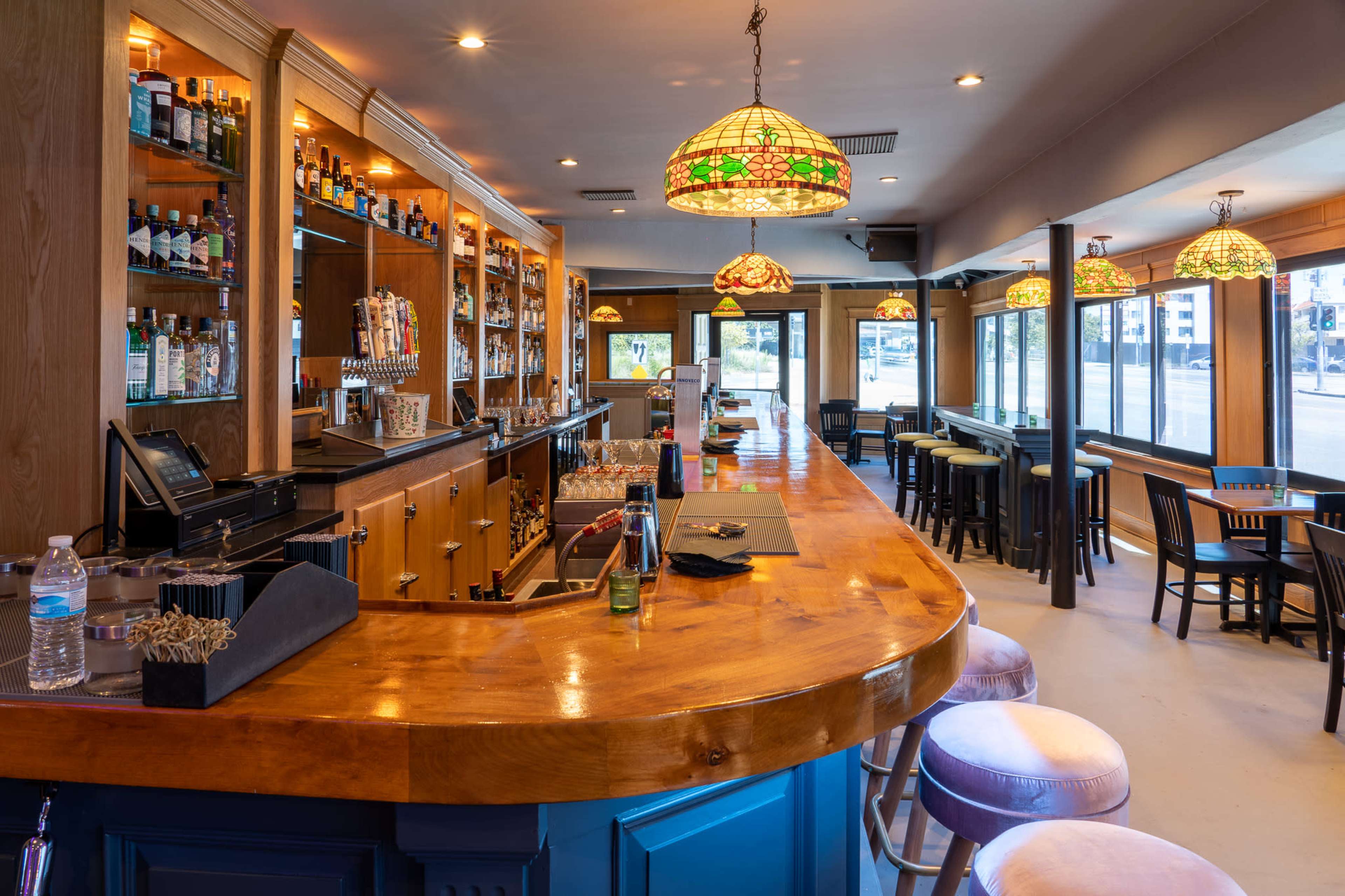 A well-lit bar area with wooden counters, shelves stocked with various bottles, and several tables and chairs arranged for patrons.