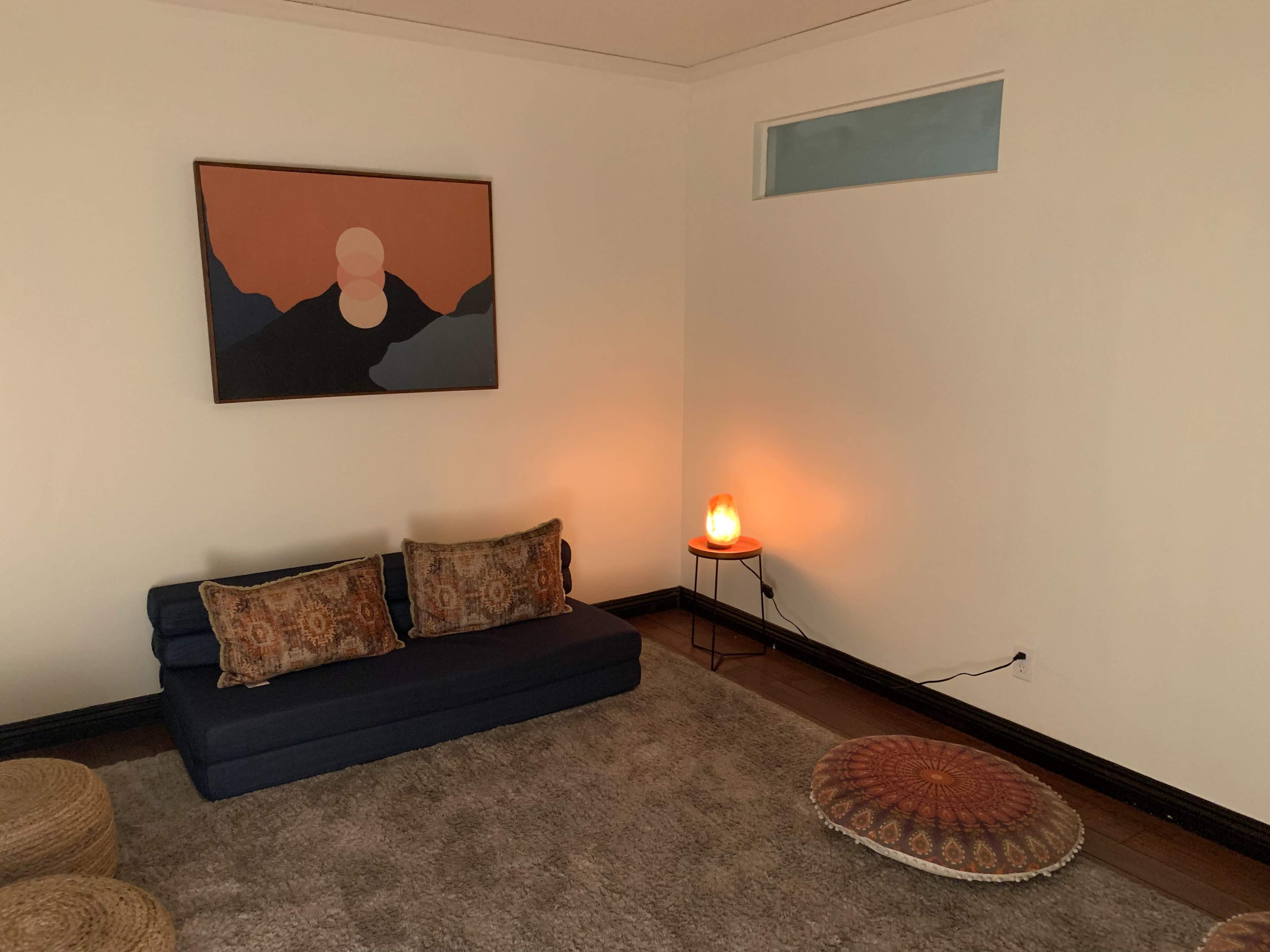 A minimalist living room features a blue sofa with patterned cushions, a round floor cushion, a decorative table with a lamp, and an abstract painting on the wall.