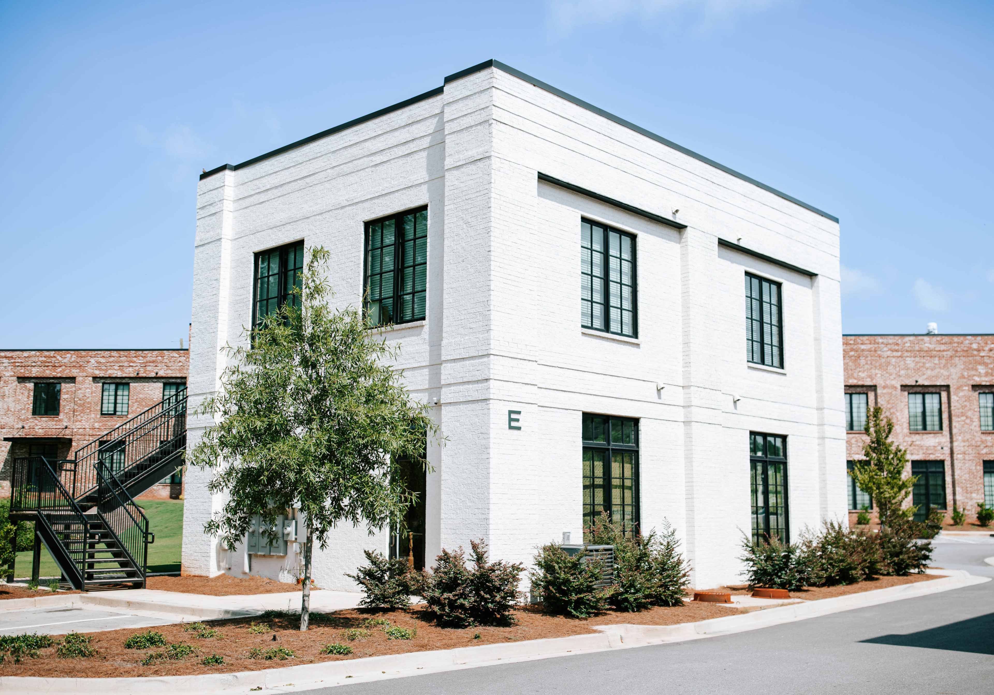 The image shows a modern two-story white brick building with large black-framed windows, surrounded by greenery and a staircase leading to the second floor.