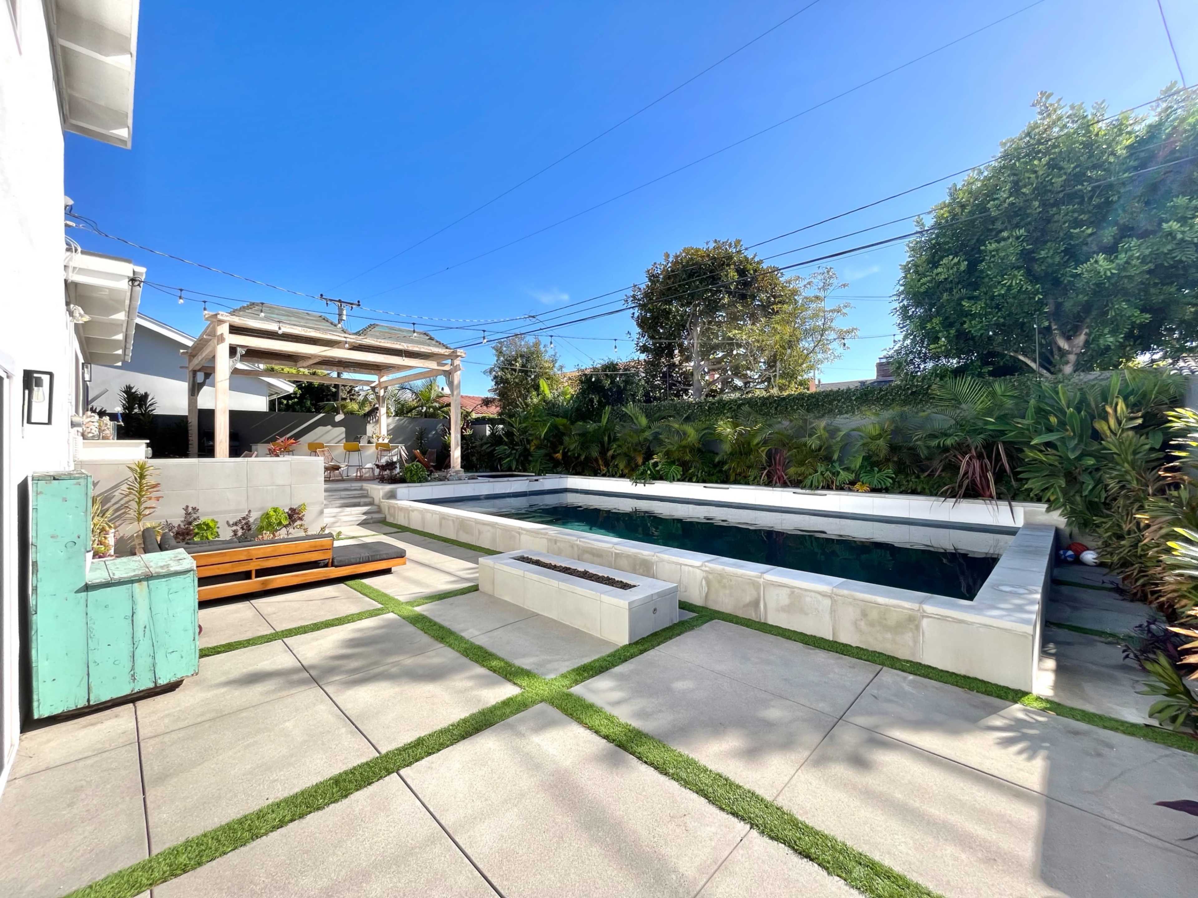 The image shows a modern backyard featuring a rectangular pool surrounded by concrete paving and lush greenery, with a wooden seating area and a covered patio.