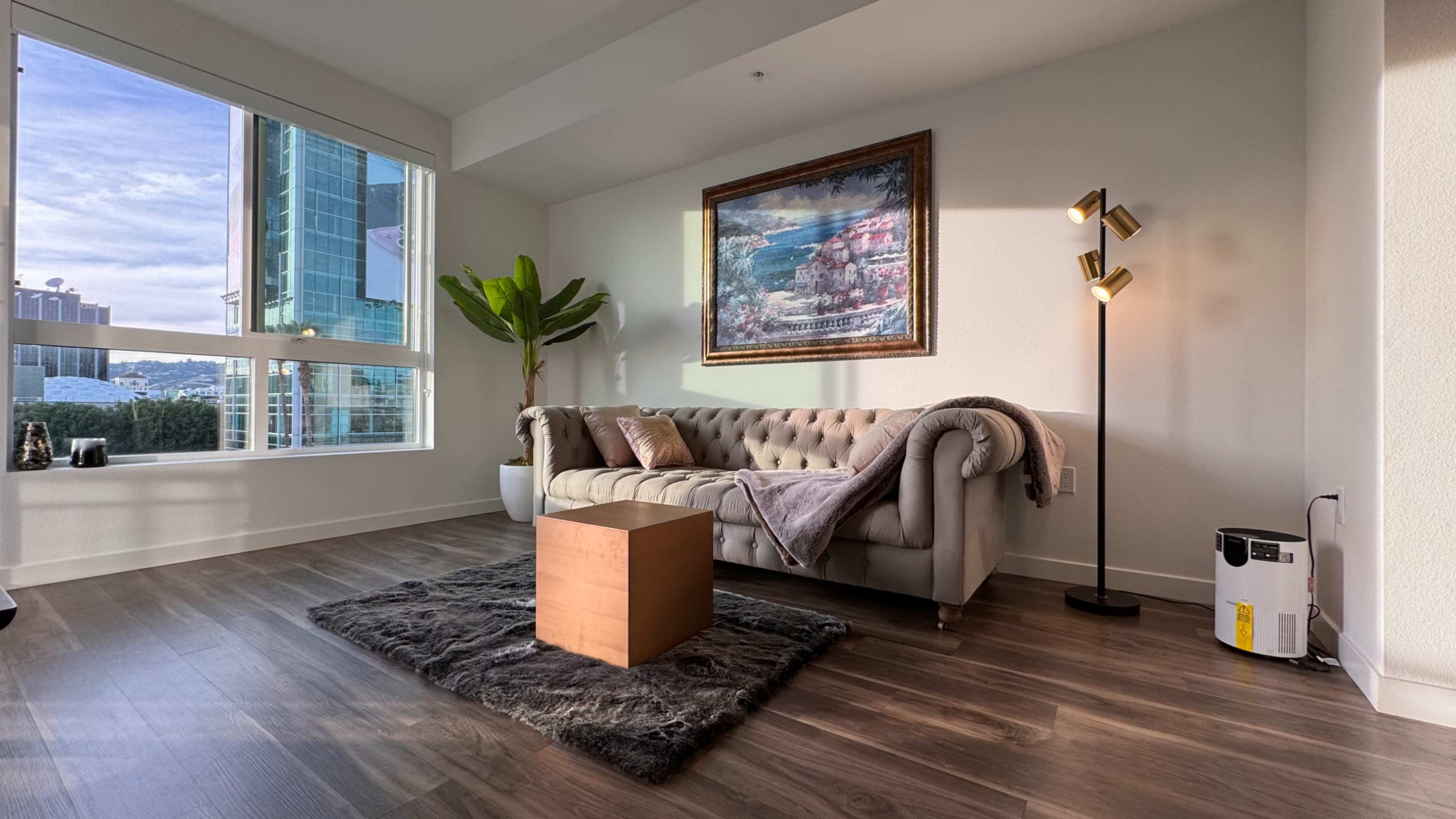 A well-lit living room featuring a beige sofa, a small wooden coffee table, a decorative painting on the wall, and a potted plant near a window.