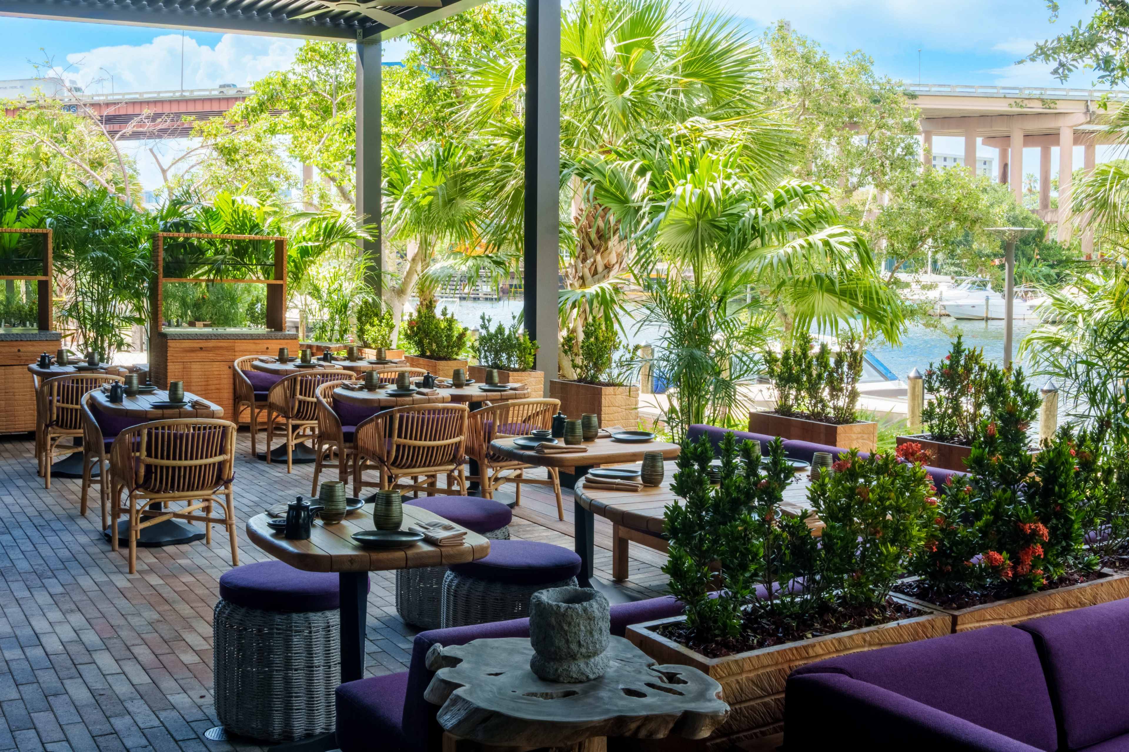 A dining area with wooden tables and chairs, surrounded by lush greenery and a waterfront view.