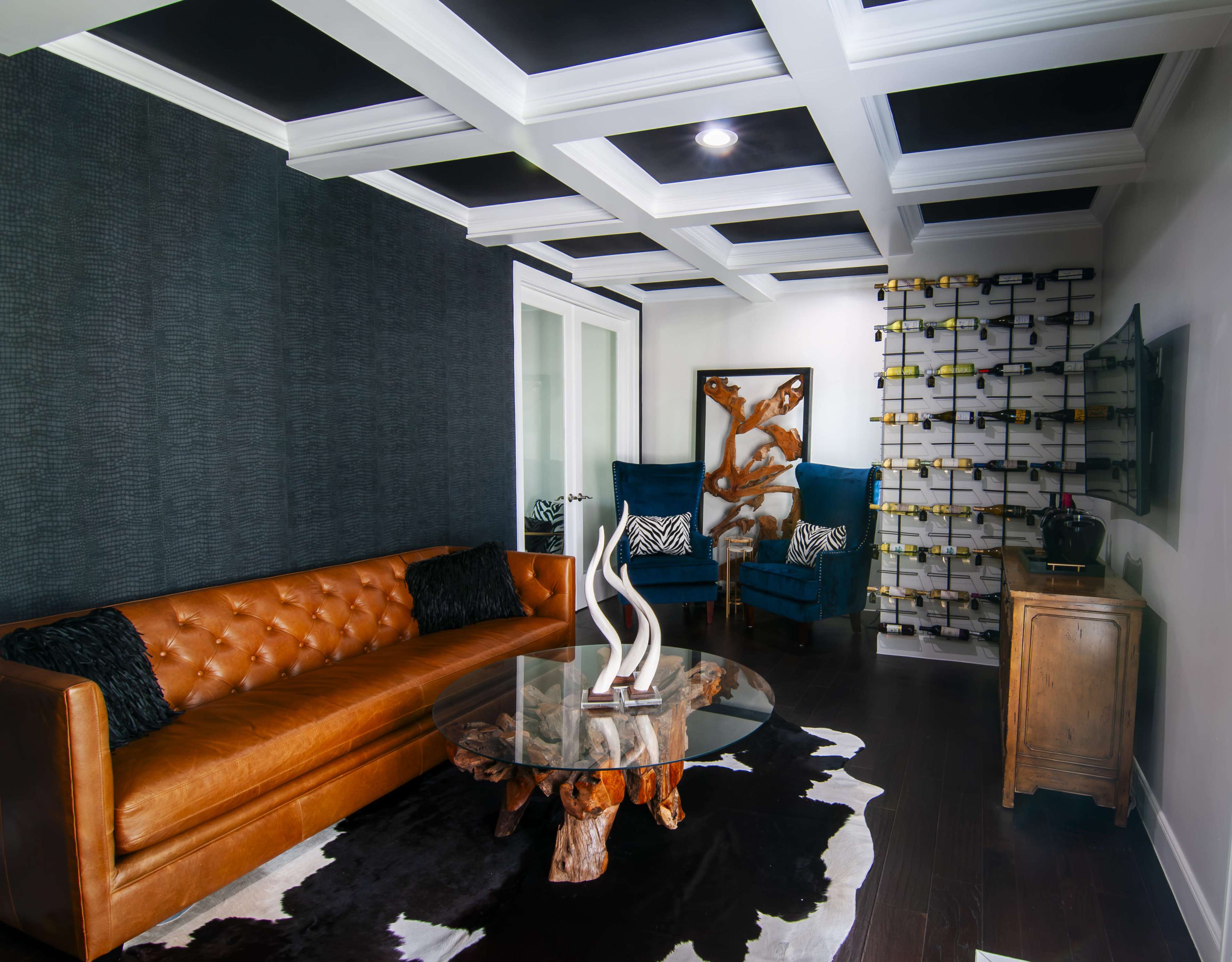 The image shows a modern living room featuring a brown tufted leather sofa, a glass coffee table on a cowhide rug, and two blue upholstered armchairs against a textured wall with a wine rack in the background.