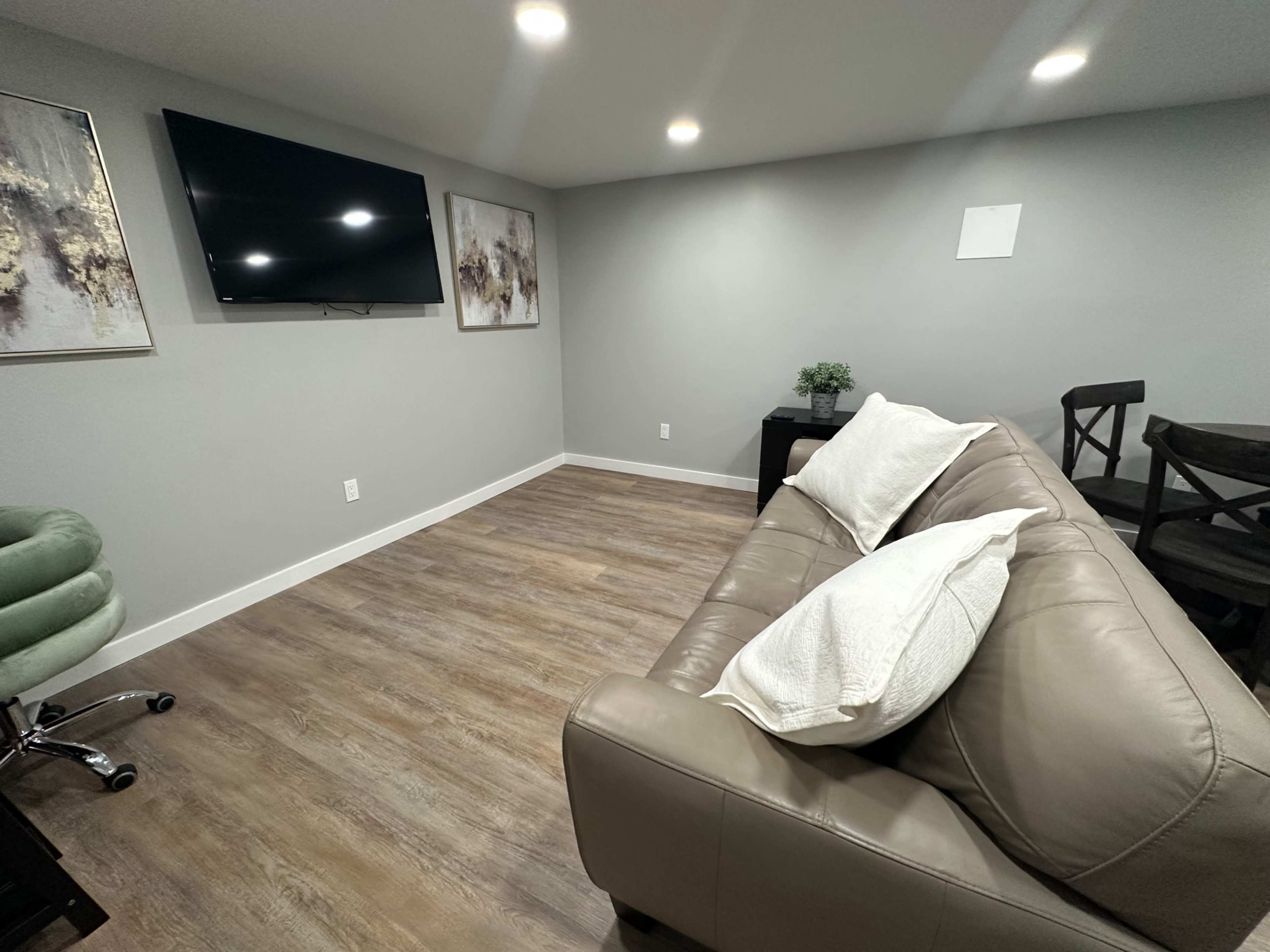 The image shows a modern, minimalistic living space with a light gray wall, a flat-screen TV mounted on the wall, a beige sofa with white cushions, and a small table beside it.