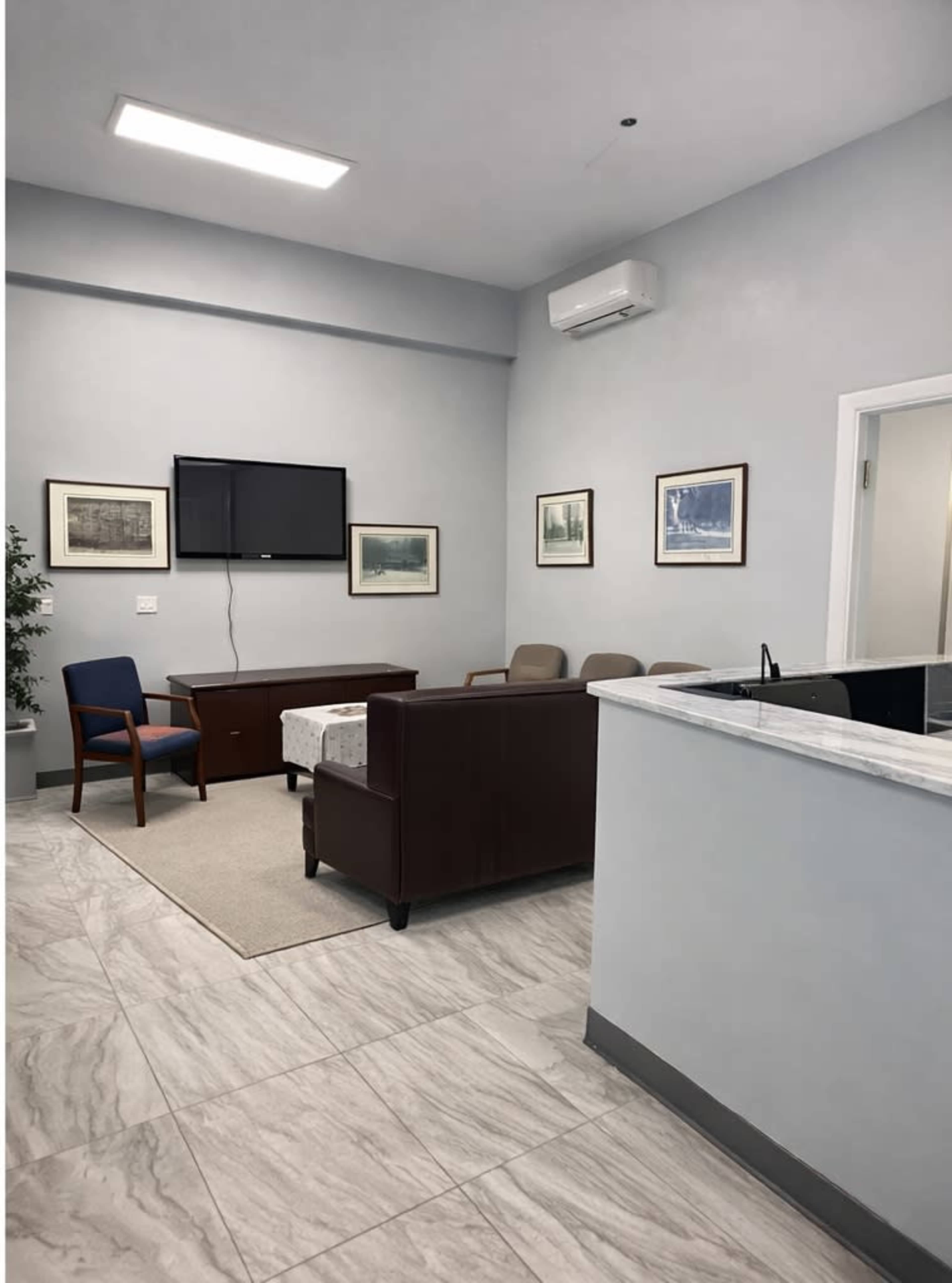 The image shows a waiting room with a sofa, chairs, and a television on the wall, along with framed artwork hanging on the gray walls.