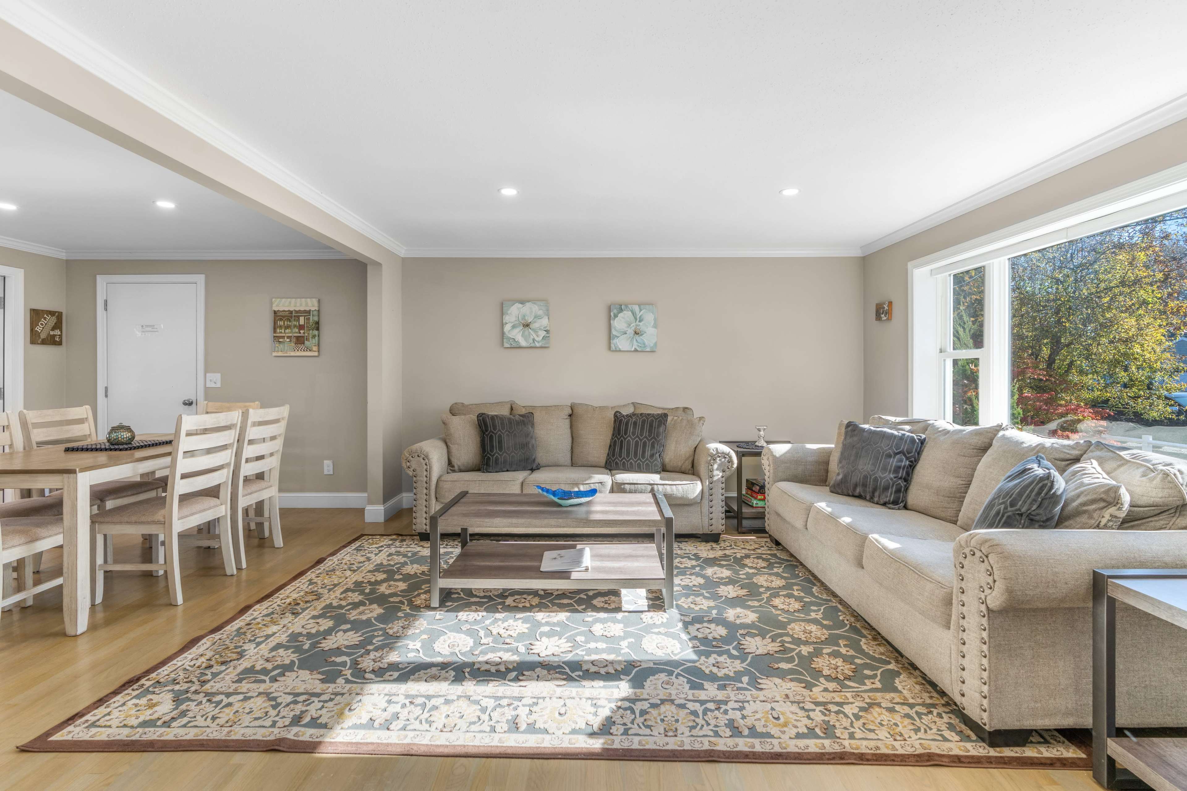 The living room features two sofas and a wooden coffee table on a patterned rug, with a dining area visible in the background.