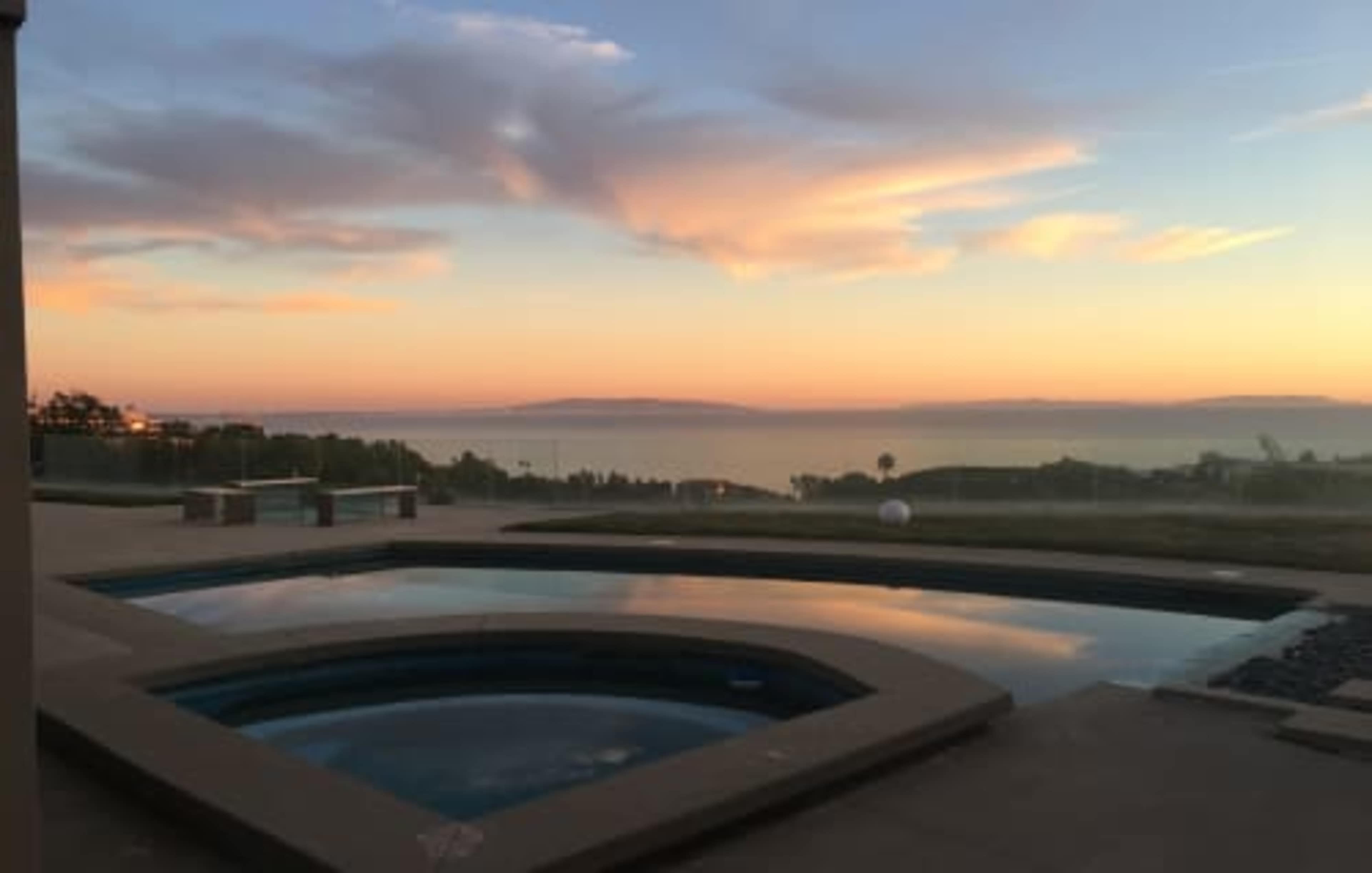 A swimming pool and hot tub overlooking a tranquil sunset with a view of the ocean and distant hills.