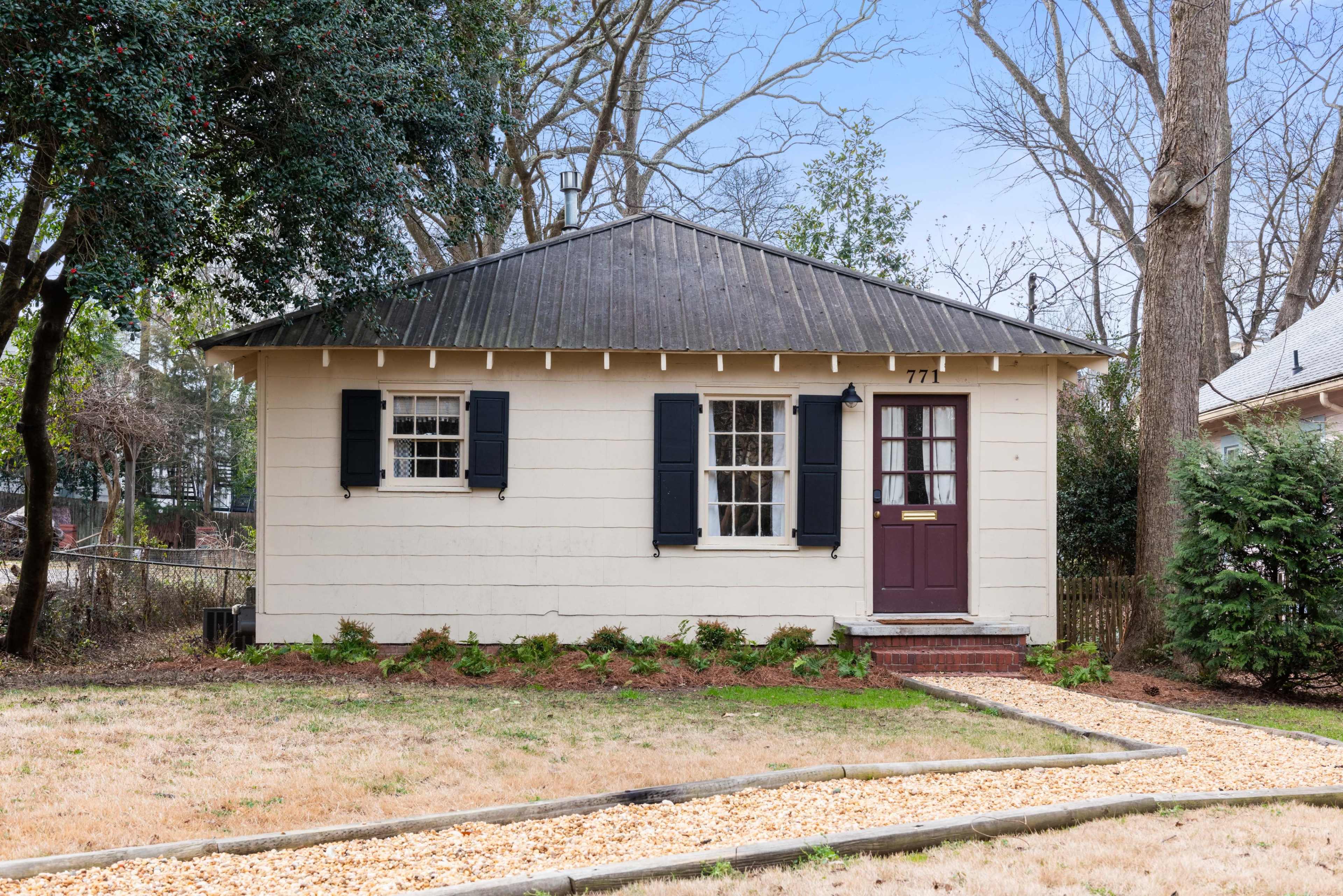 The image shows a single-story house with a gabled roof, black shutters, and a pathway lined with gravel leading up to the front door.