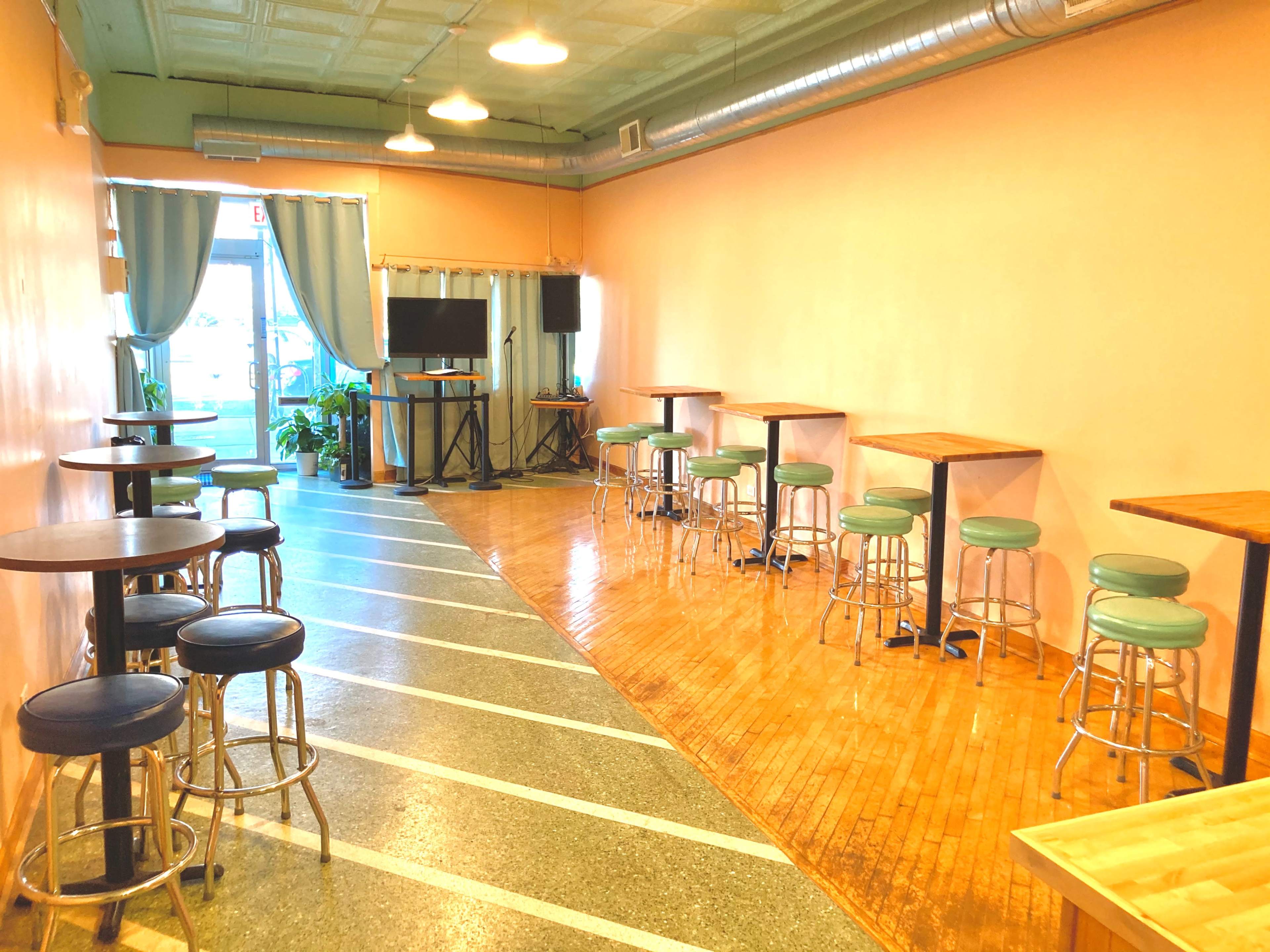 A spacious room with wooden floors, a row of tall tables and stools on one side, and large windows letting in natural light.