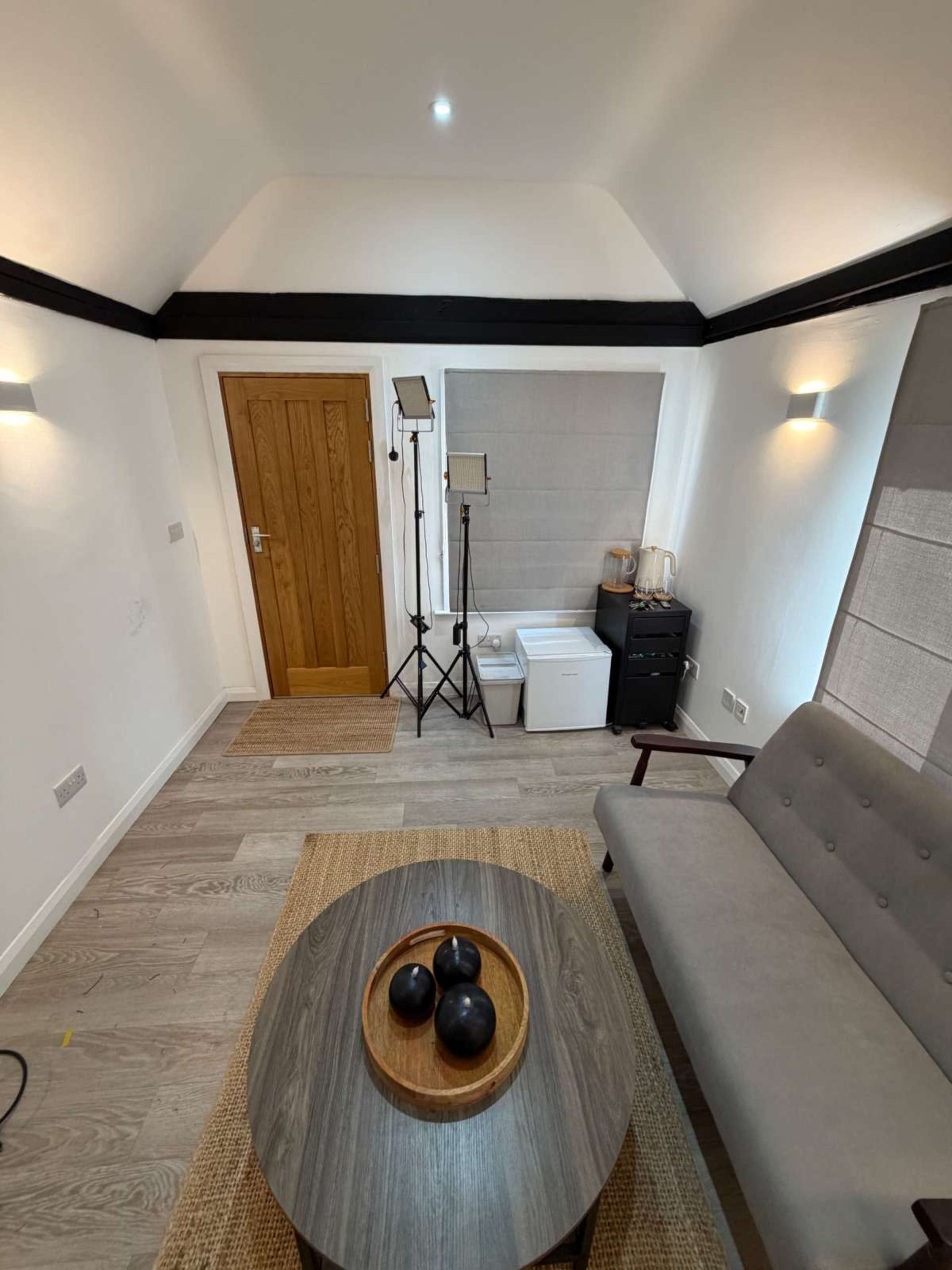 The image shows a minimally furnished room with a gray sofa, a round wooden coffee table with three black decor pieces, and a tall tripod with lights in one corner.
