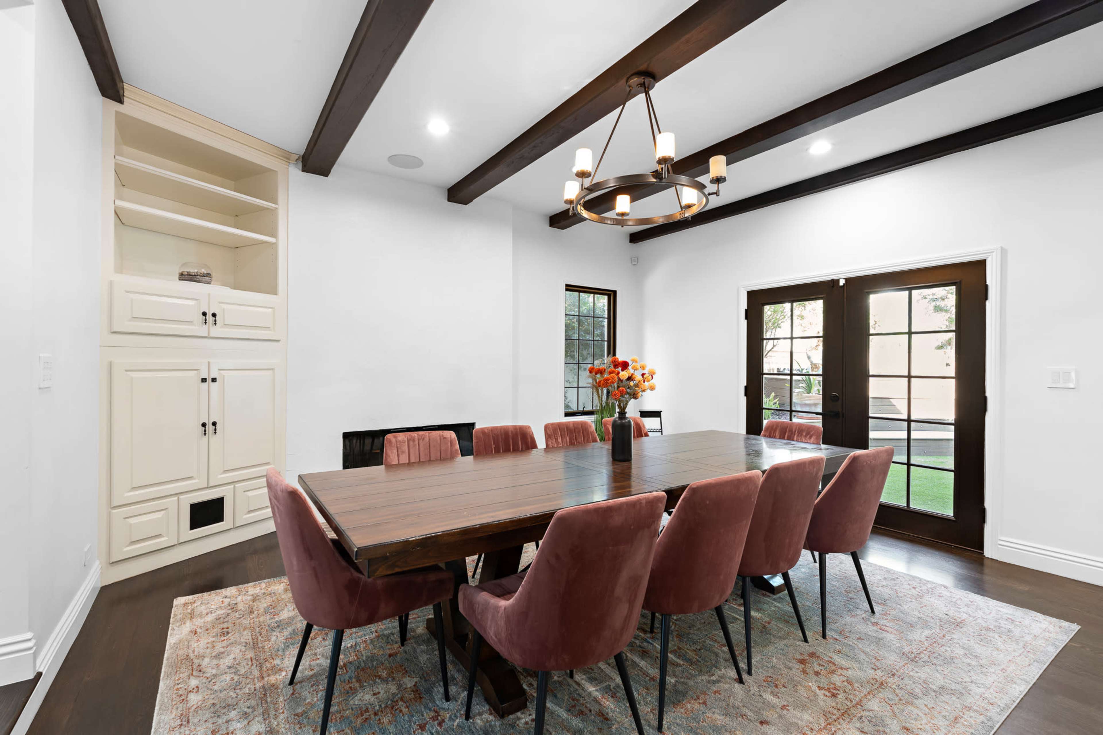 A dining room features a large wooden table surrounded by pink upholstered chairs, with a chandelier overhead and a view of an outdoor area through glass doors.