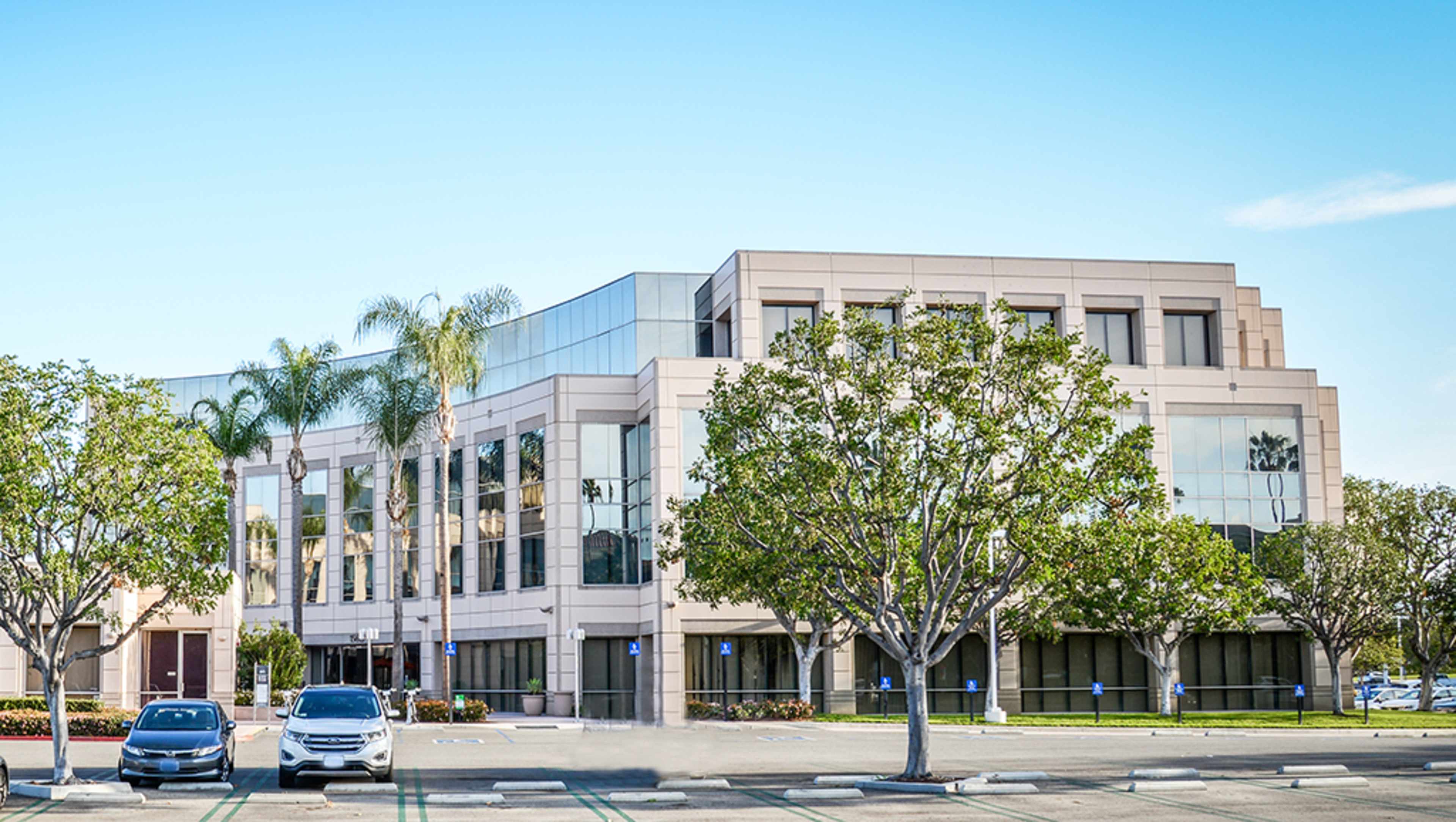 A modern office building with large windows is surrounded by landscaped trees and parked cars in the foreground.