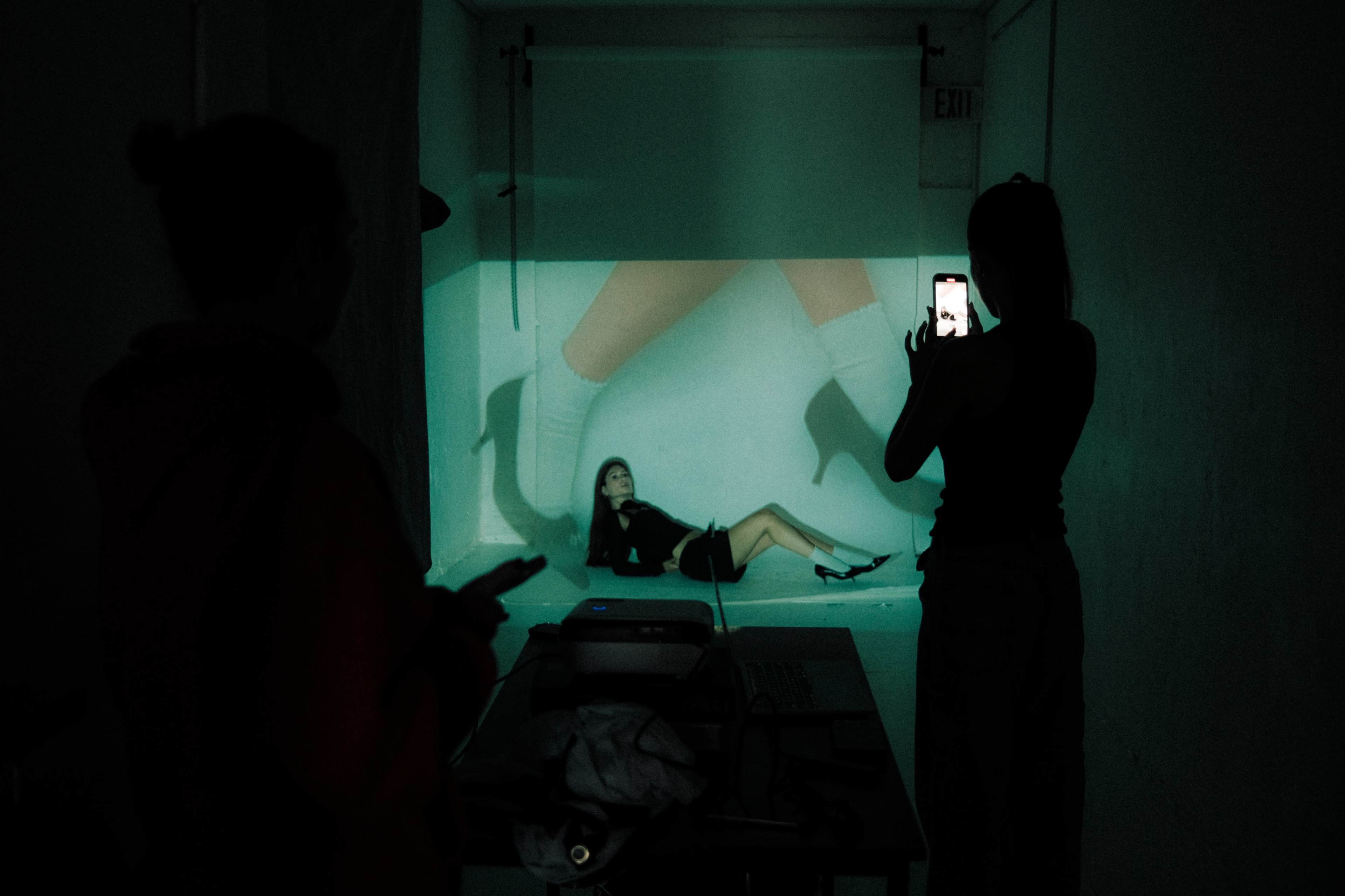 A woman poses on the ground in front of a projected image of legs, while another person takes a photo of her with a smartphone.