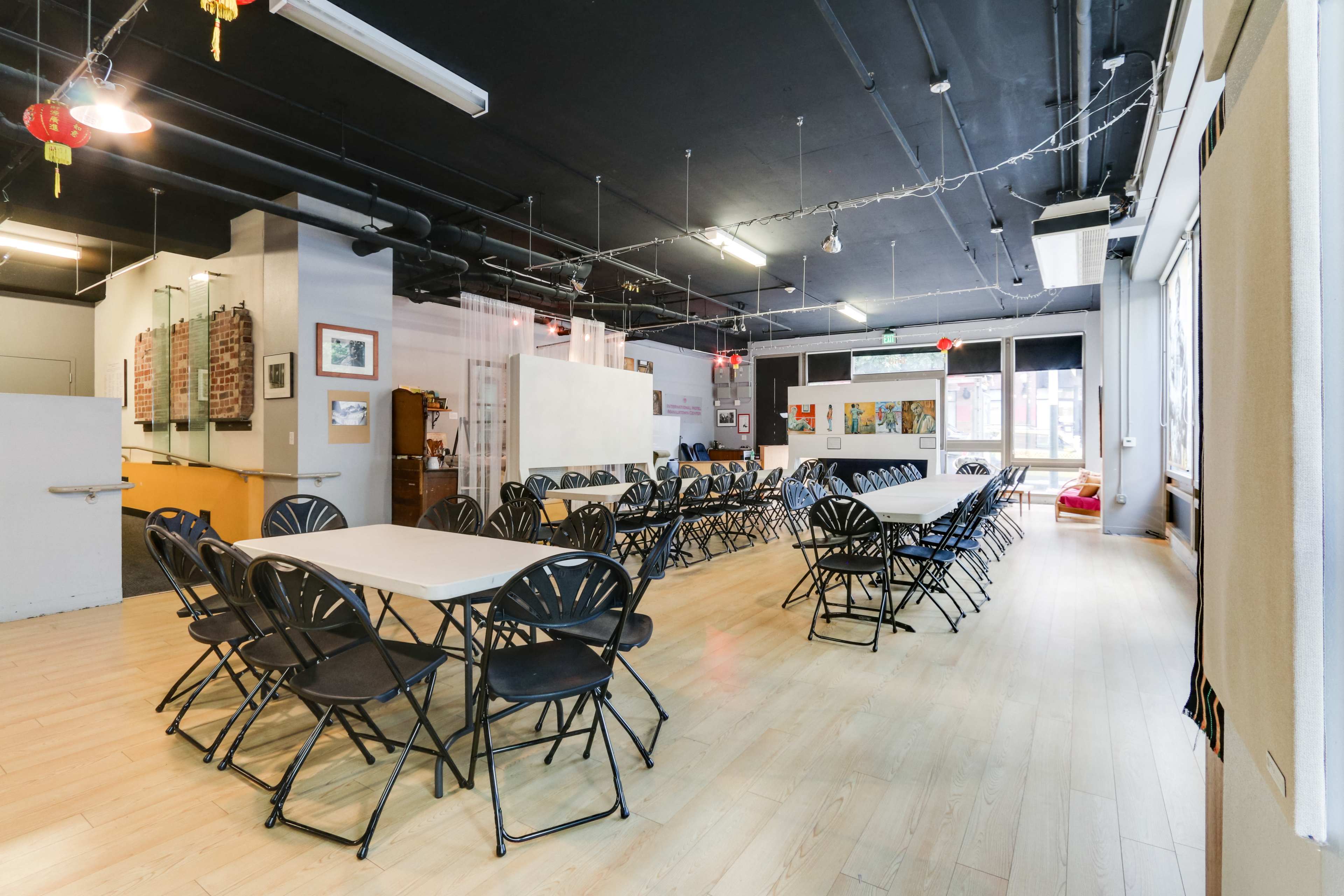 A spacious room with multiple long tables and black folding chairs arranged in rows, illuminated by overhead lights and featuring large windows.