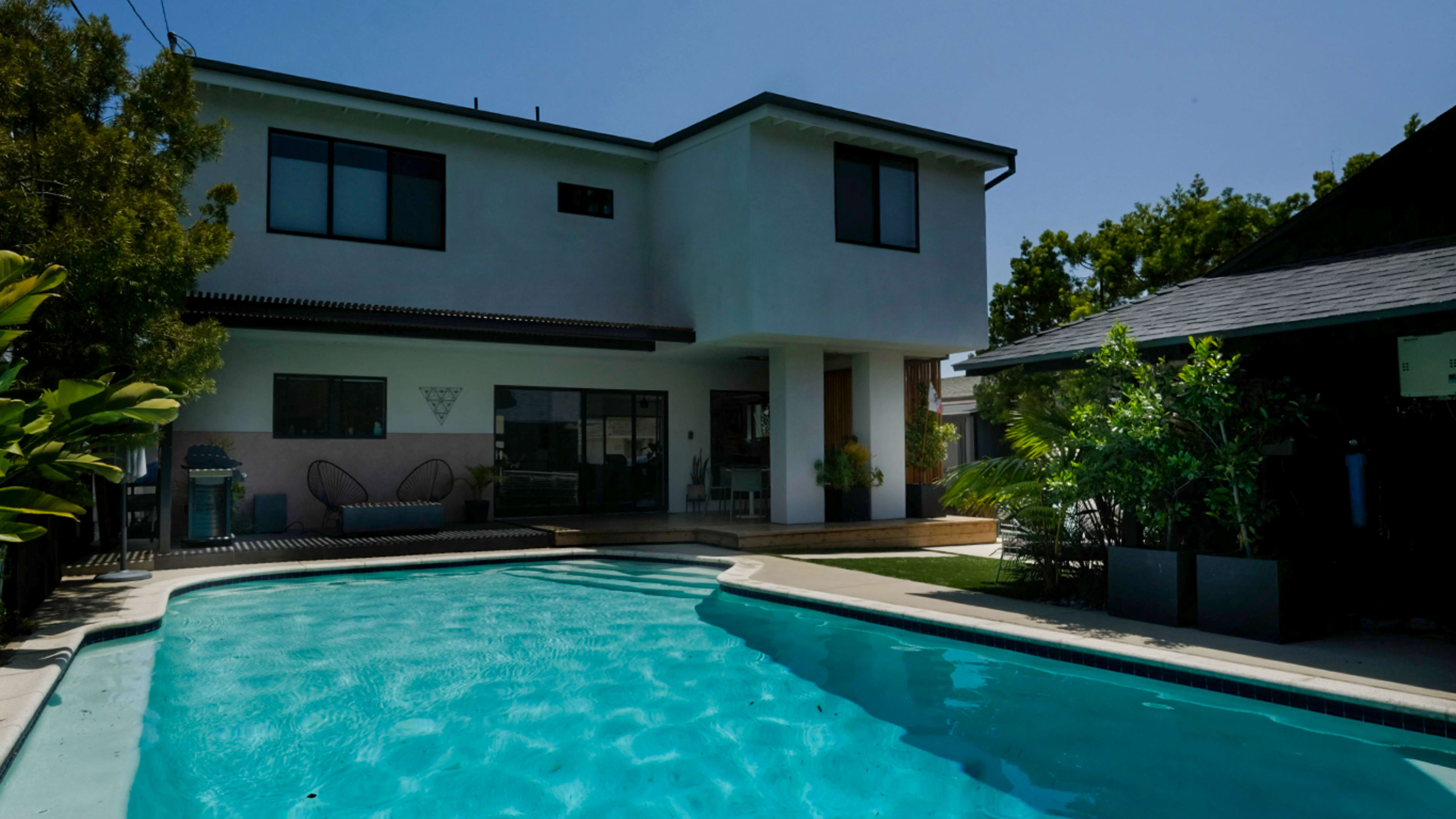 A modern two-story house with large windows, a swimming pool in the foreground, and landscaped greenery surrounding the area.