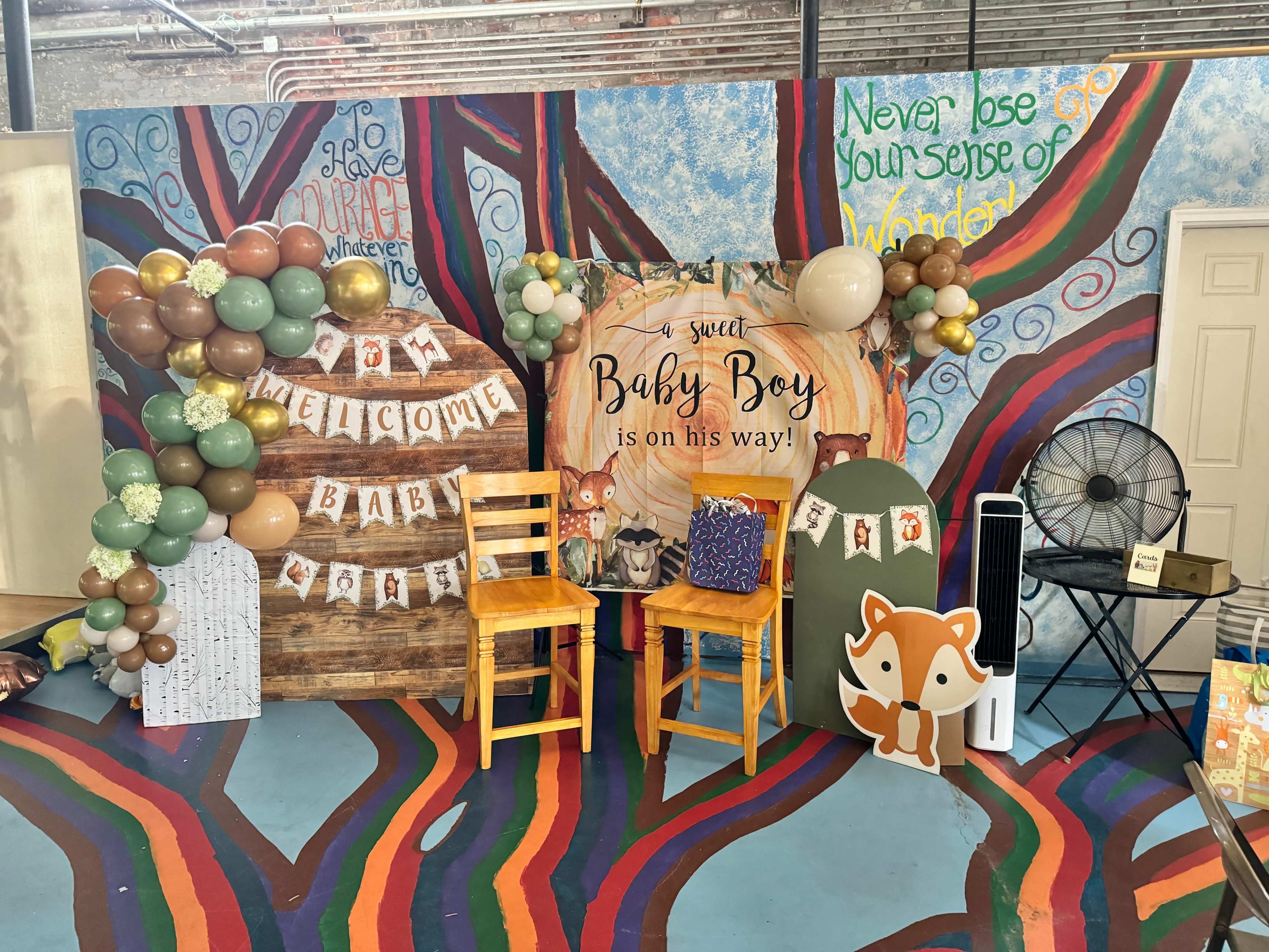 A decorated event space featuring two wooden chairs and a backdrop with a whimsical theme of balloons, a sign that reads "Welcome Baby," and a playful fox graphic.