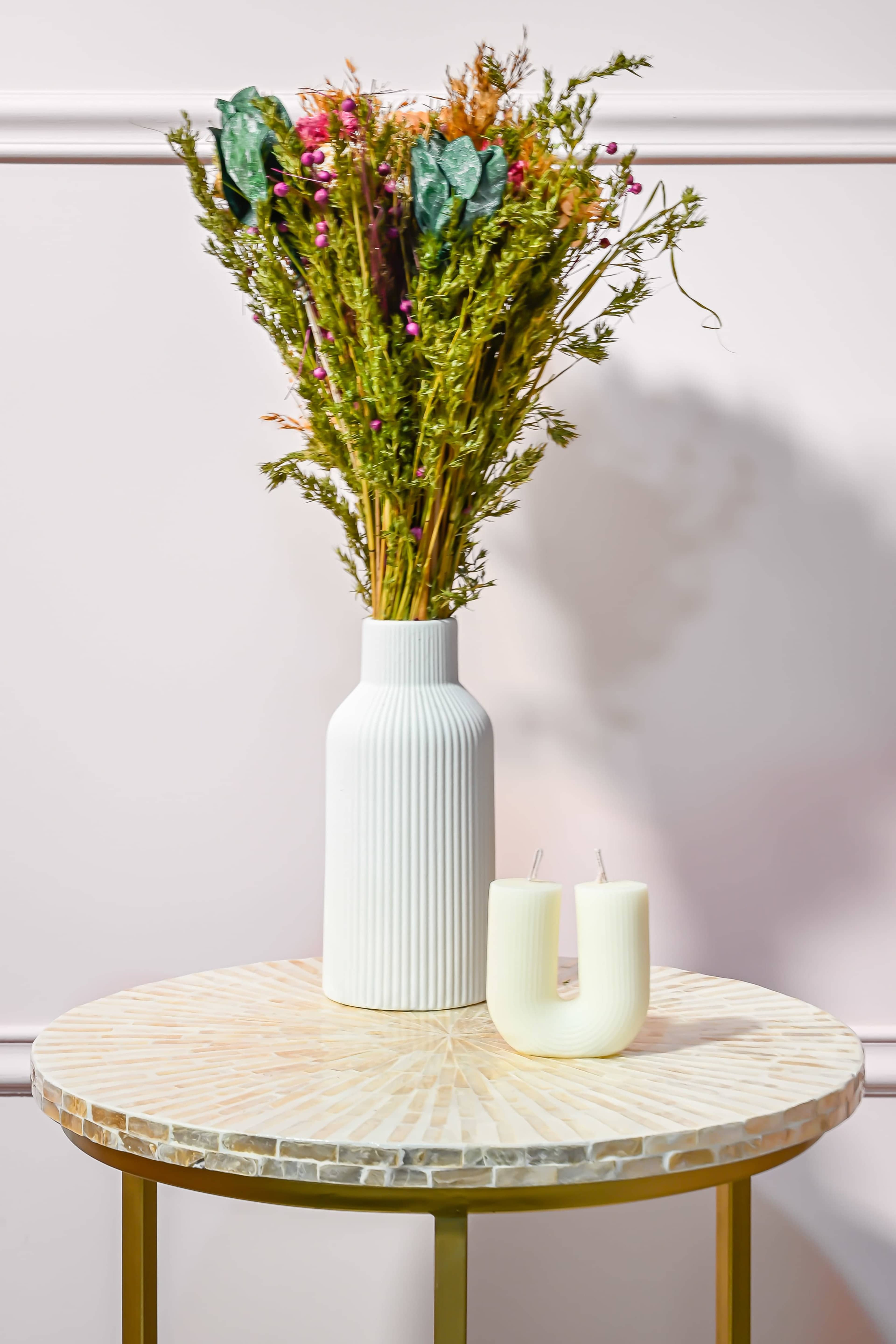 A white vase filled with dried flowers is positioned on a round table with a textured surface, accompanied by two white candles.
