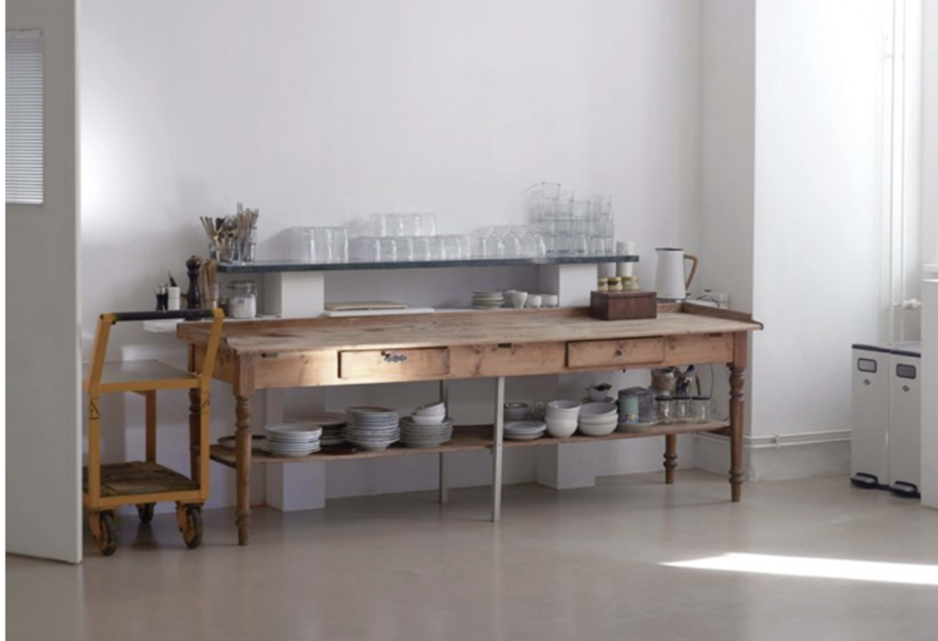 A wooden table with a drawer and a shelf above it holds glassware and plates in a bright, minimalist room.