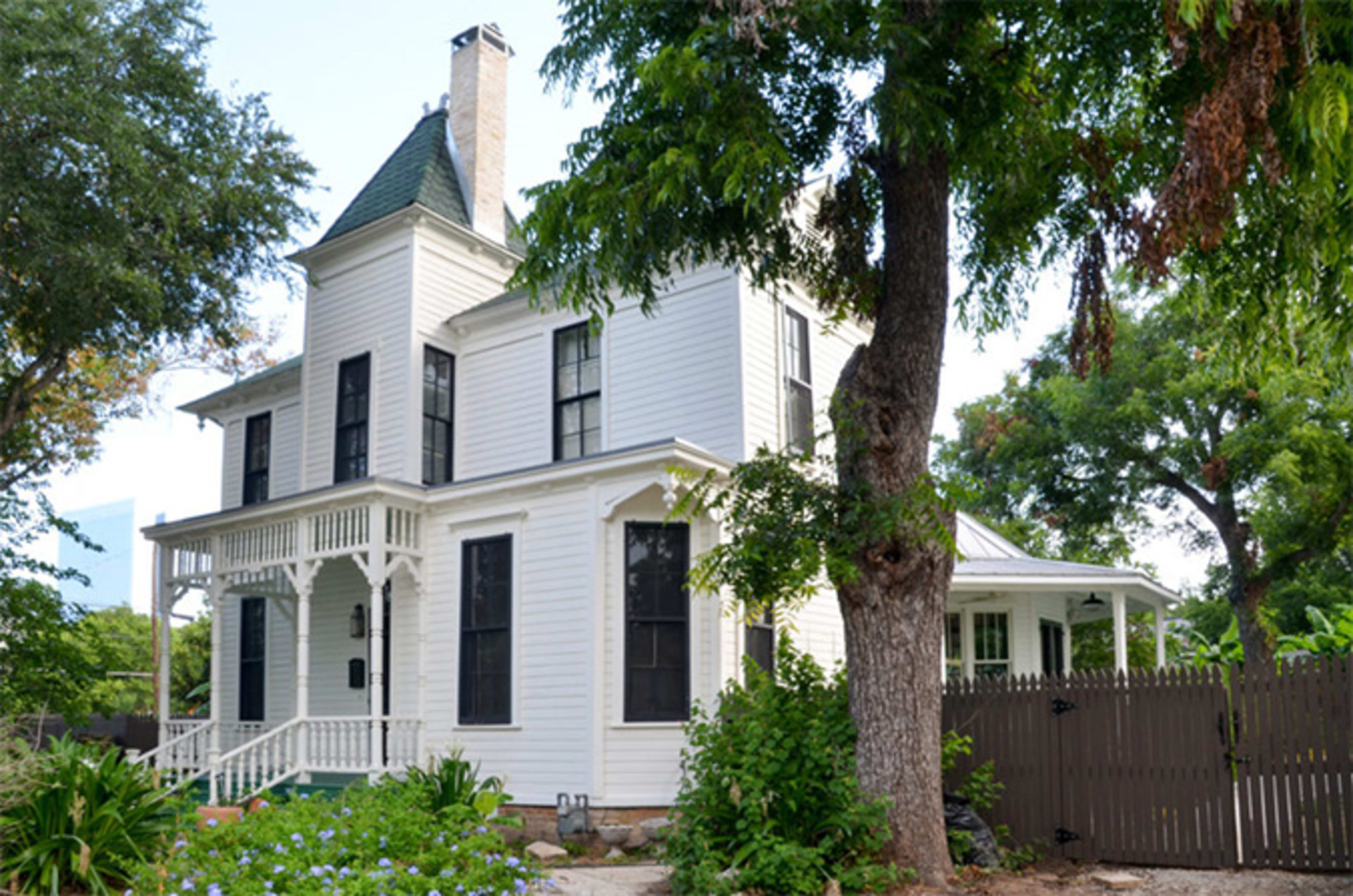 The image shows a two-story white Victorian-style house surrounded by green foliage and a wooden fence.