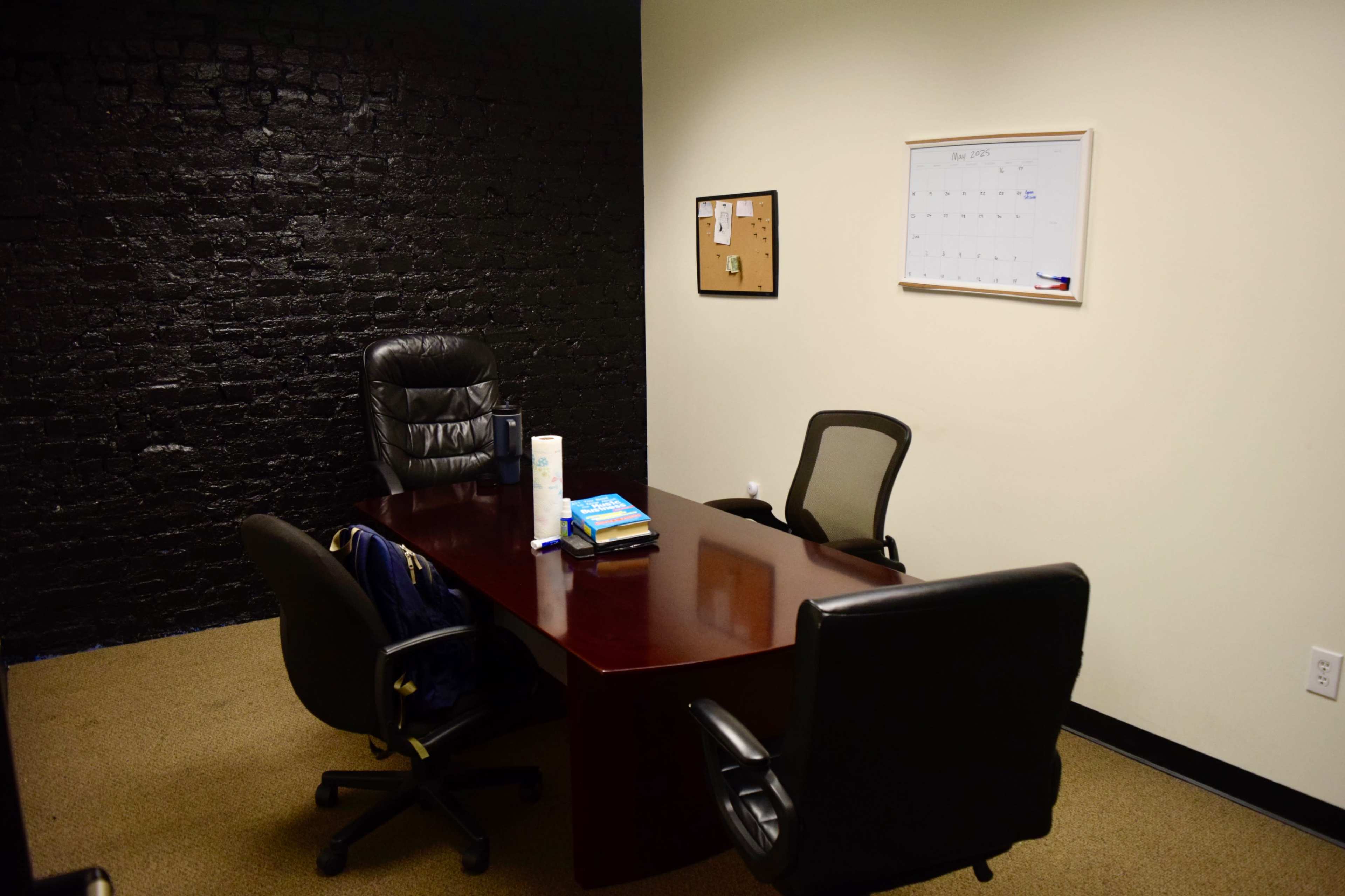 The image shows a small office space with a dark brick wall, a wooden conference table, two black office chairs, a calendar on the wall, and a corkboard.