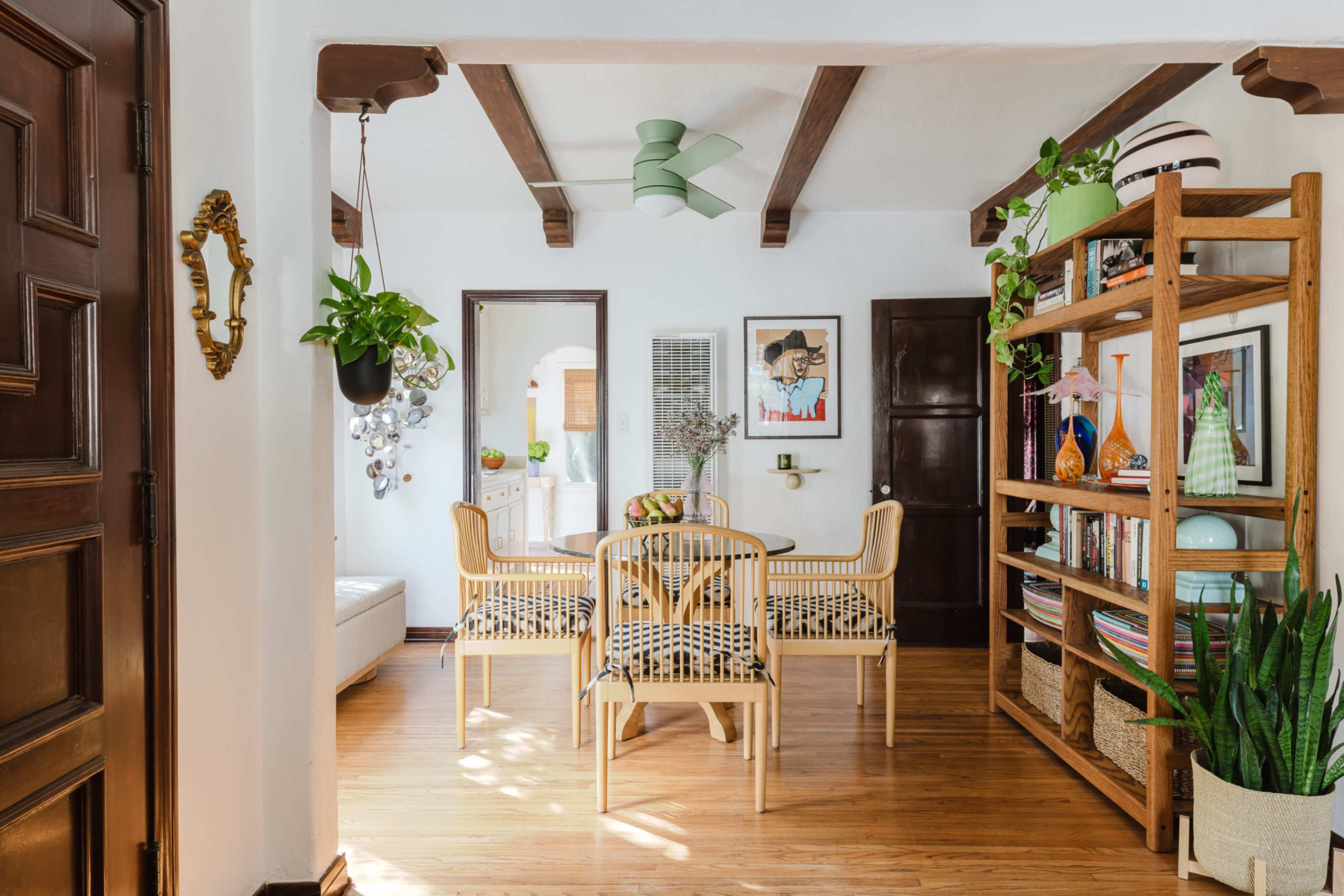 The image shows a bright dining area with wooden beams, a round table surrounded by chairs, and a decorative shelf filled with plants and colorful items.