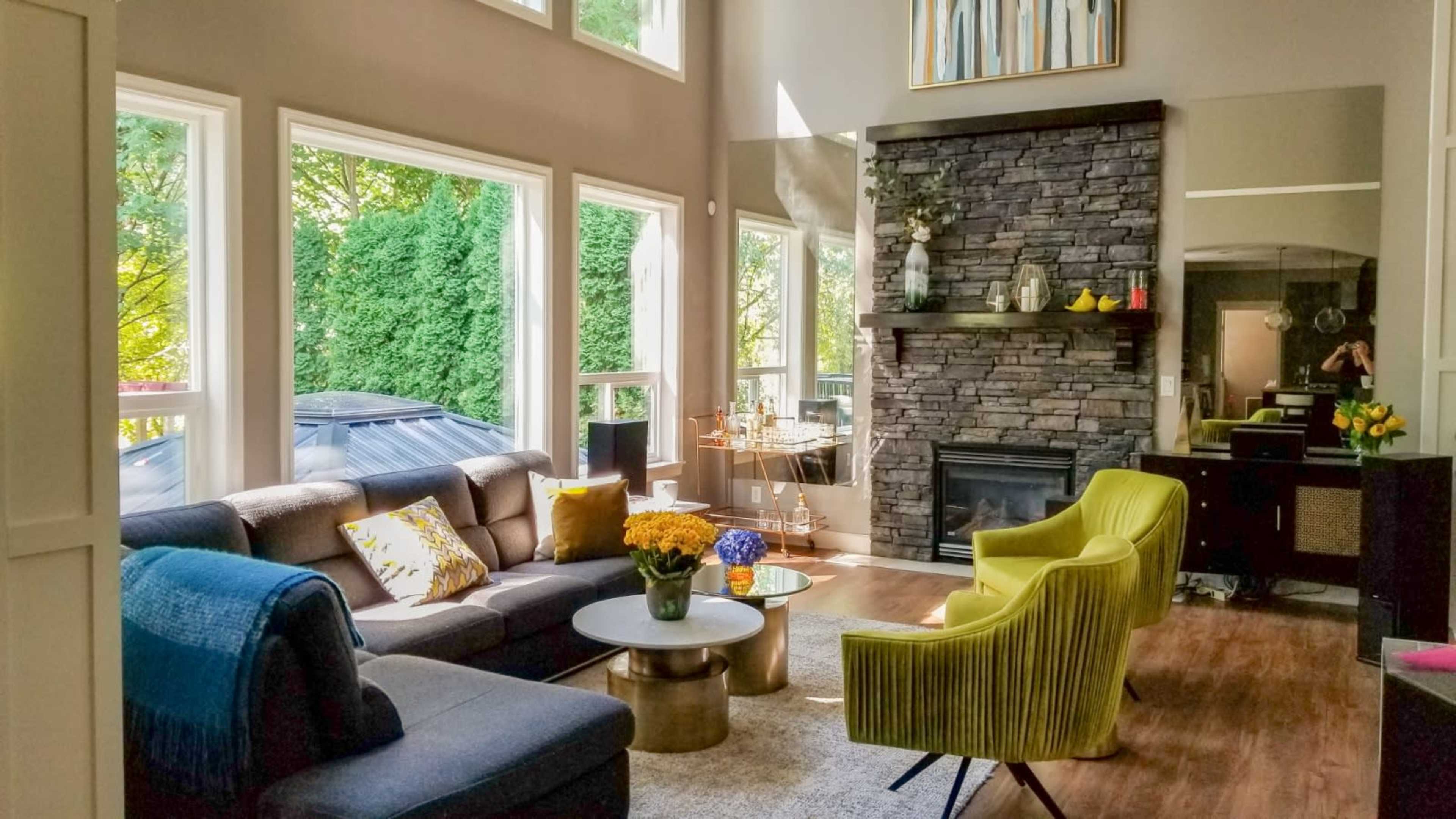 A modern living room with large windows, a stone fireplace, a circular coffee table, and bright yellow accent chairs.