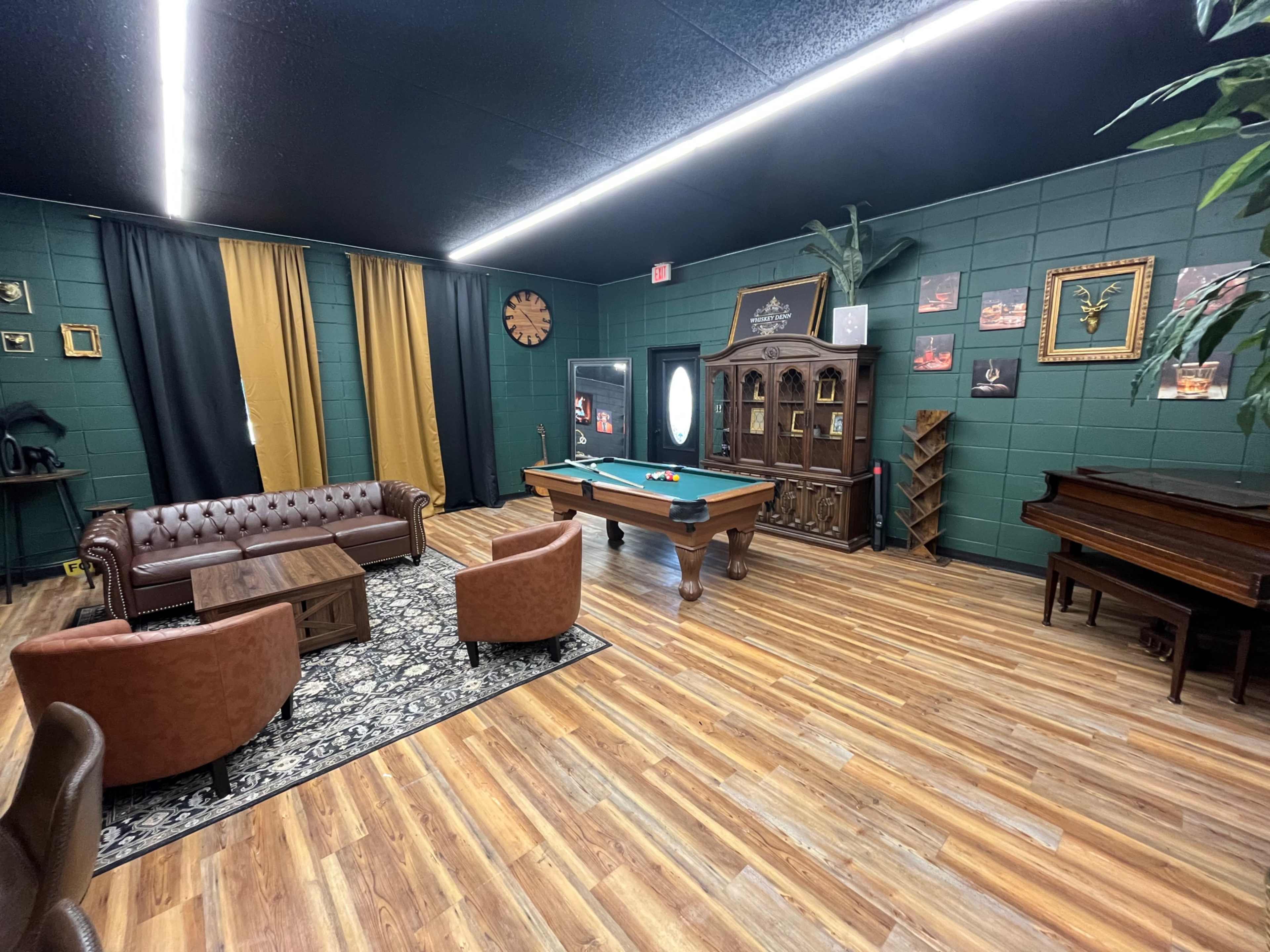 The image shows a stylish lounge area featuring a pool table, a brown leather sofa, wooden chairs, a piano, and decorative plants against a green wall.
