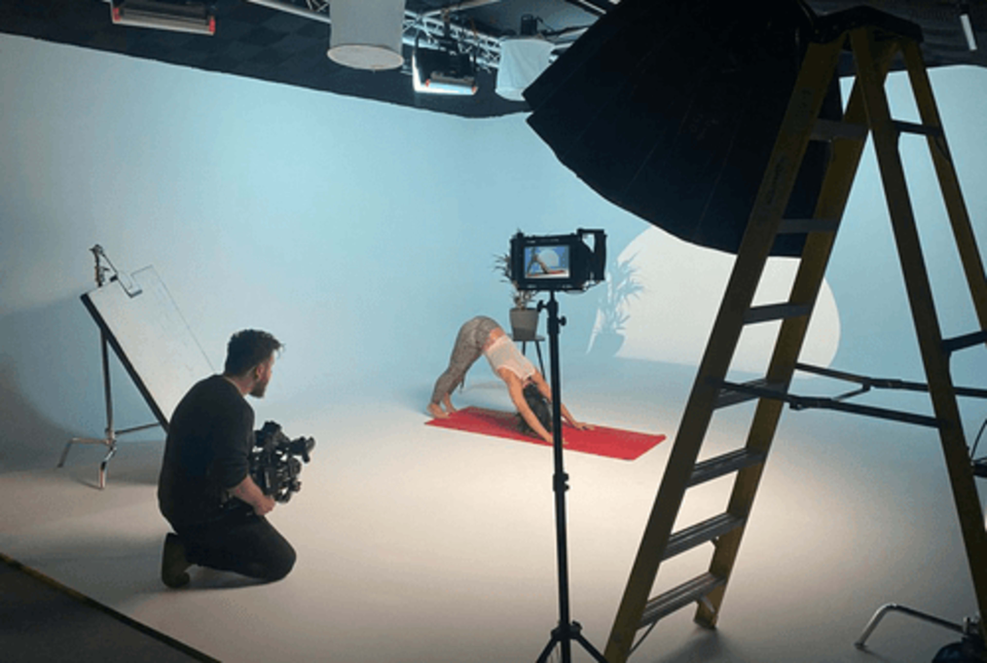 A person is practicing yoga on a red mat while another person sets up a camera on a tripod nearby in a studio with a light blue backdrop.