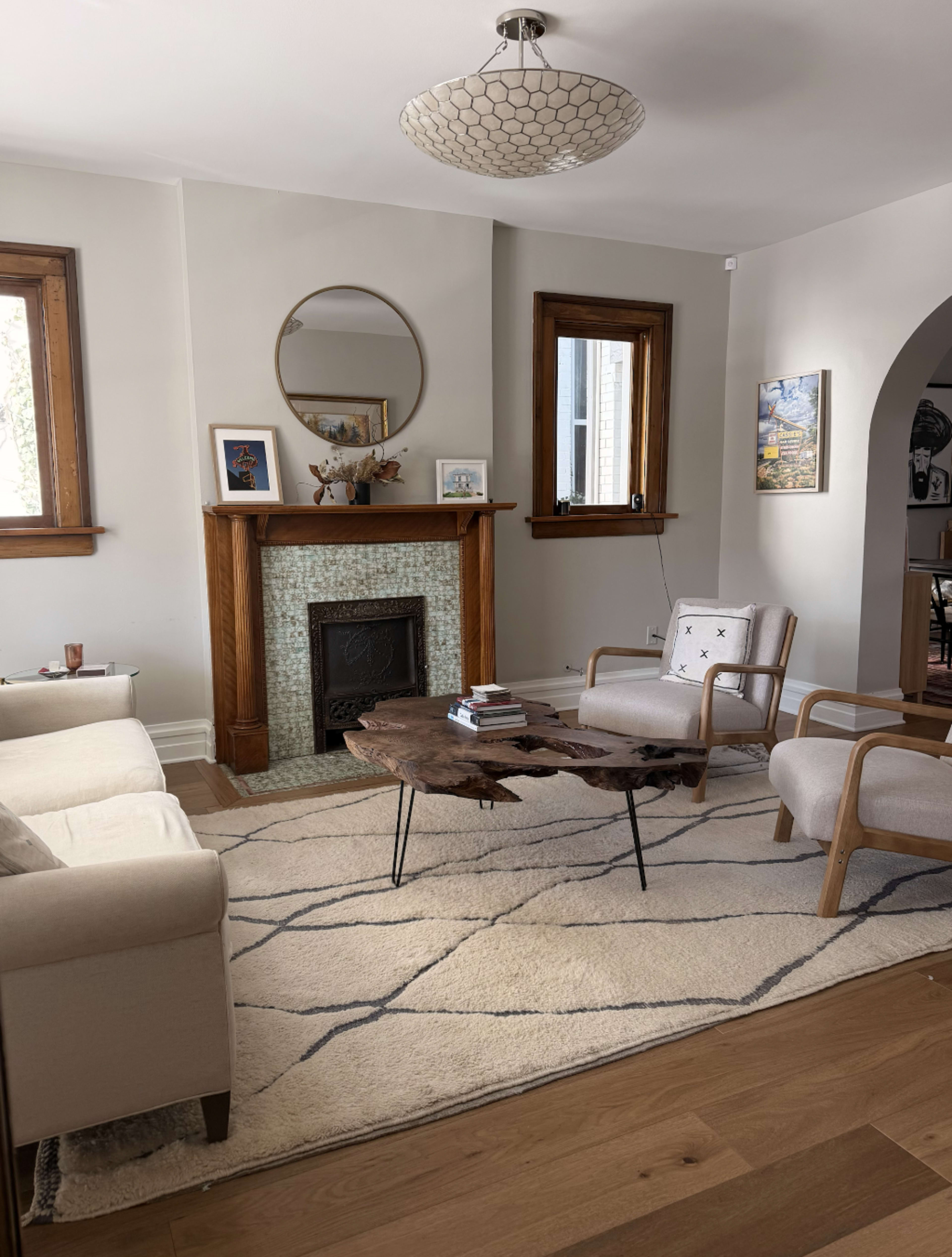 The image shows a living room featuring a fireplace with a circular mirror above it, a unique wooden coffee table, two armchairs, and a light-colored sofa, all arranged on a patterned rug.