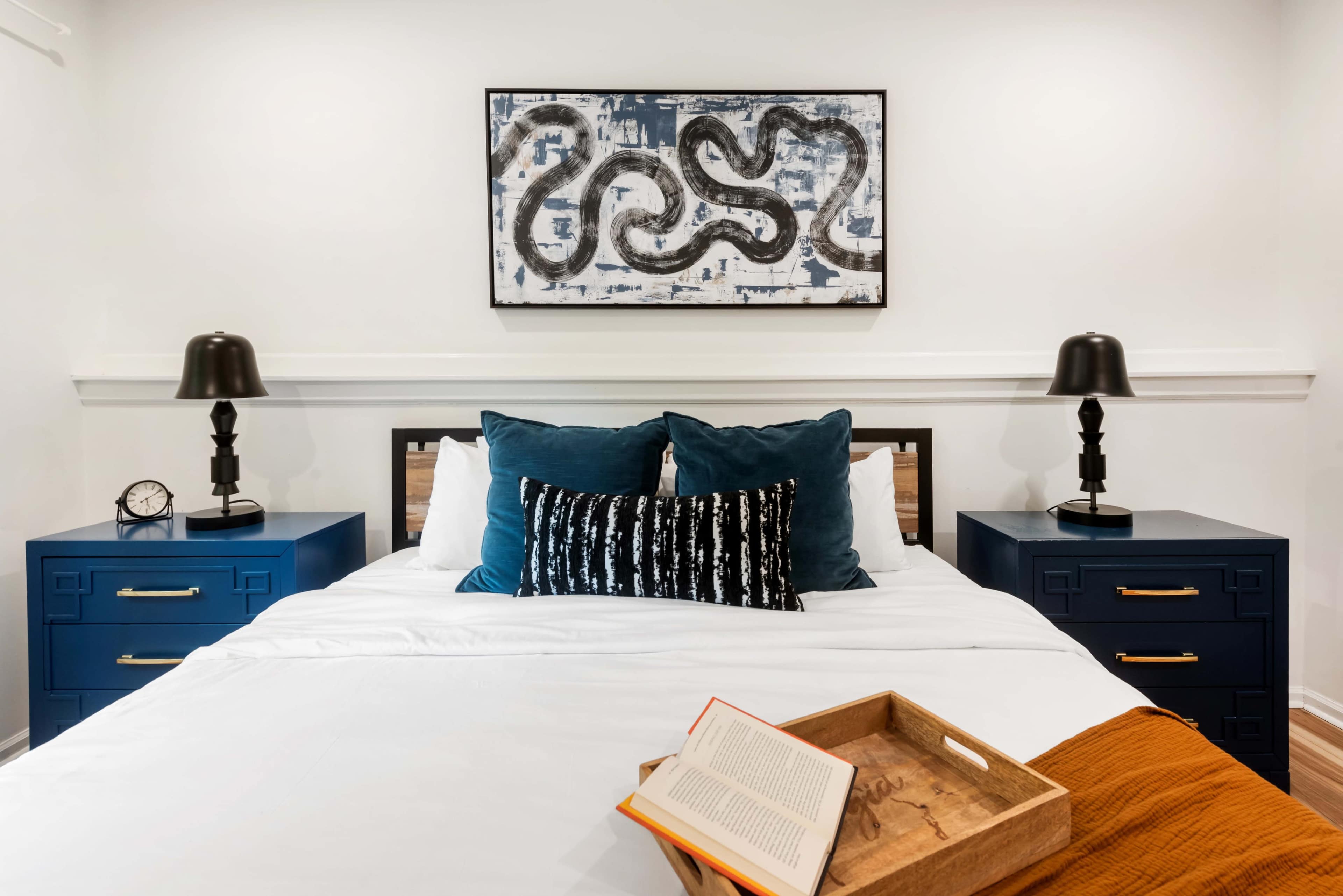 The image shows a neatly arranged bedroom with a central bed, two nightstands with lamps, and a piece of abstract artwork above the headboard.