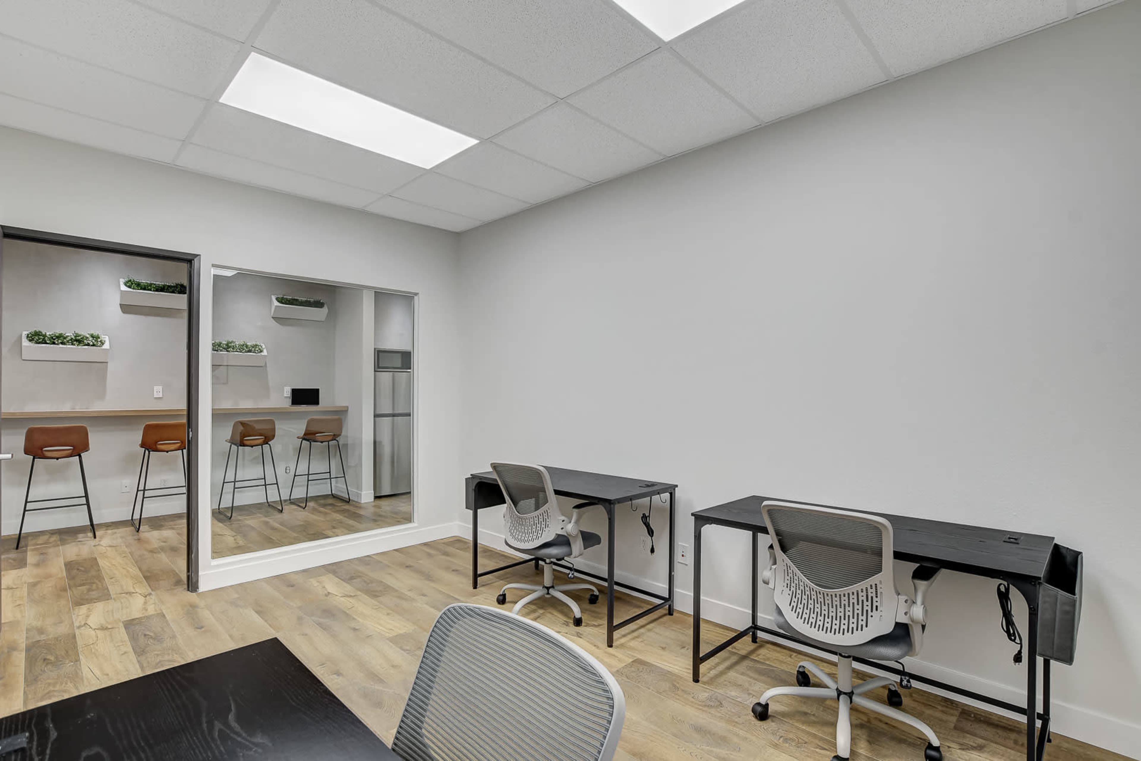 A modern office space with two black desks, gray office chairs, and a bar area with three stools against a mirrored wall.