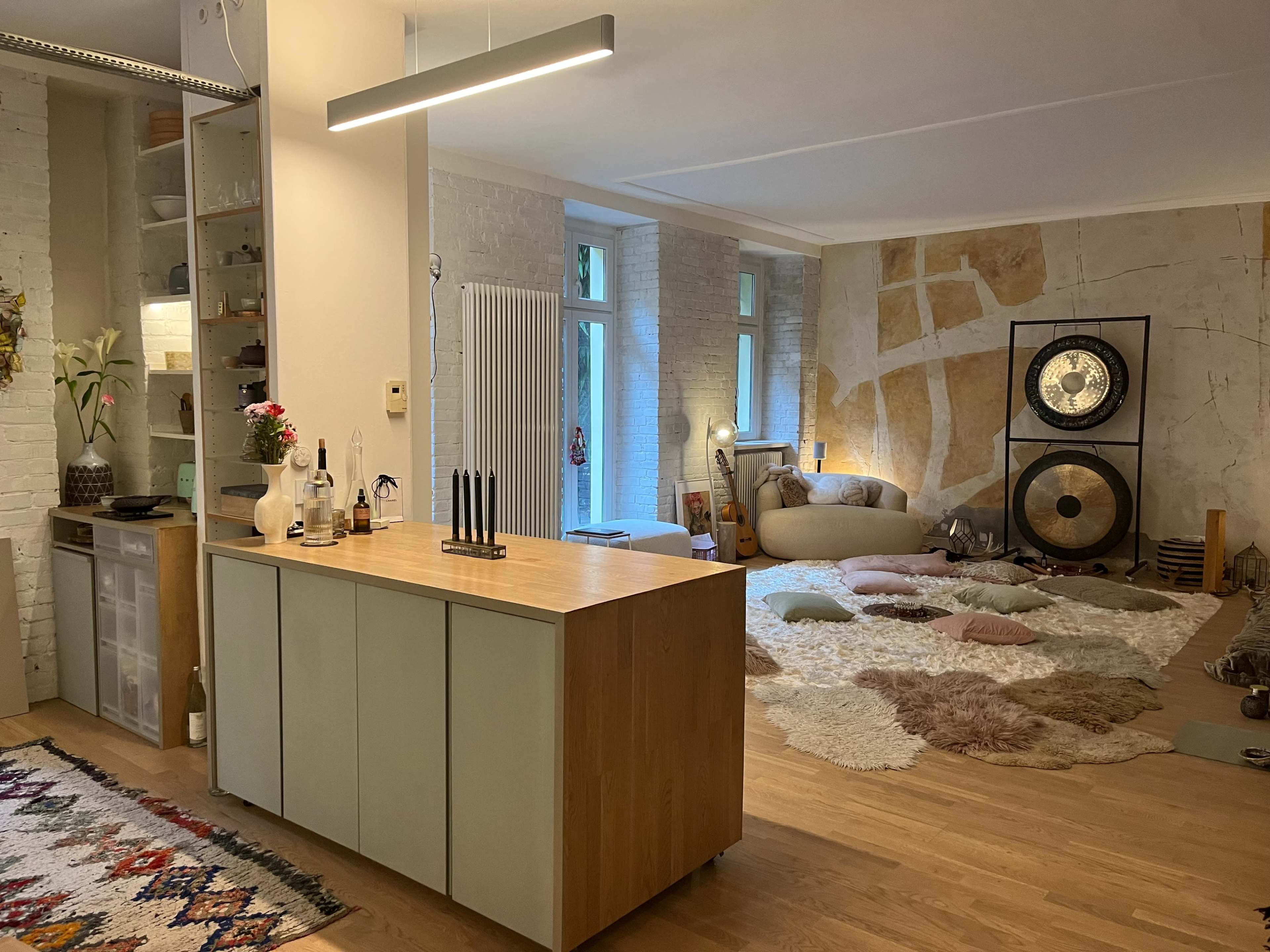 The image shows a modern, open-concept living space featuring a kitchen island with a wooden top, a cozy seating area with multiple rugs, and large windows allowing natural light to filter in.