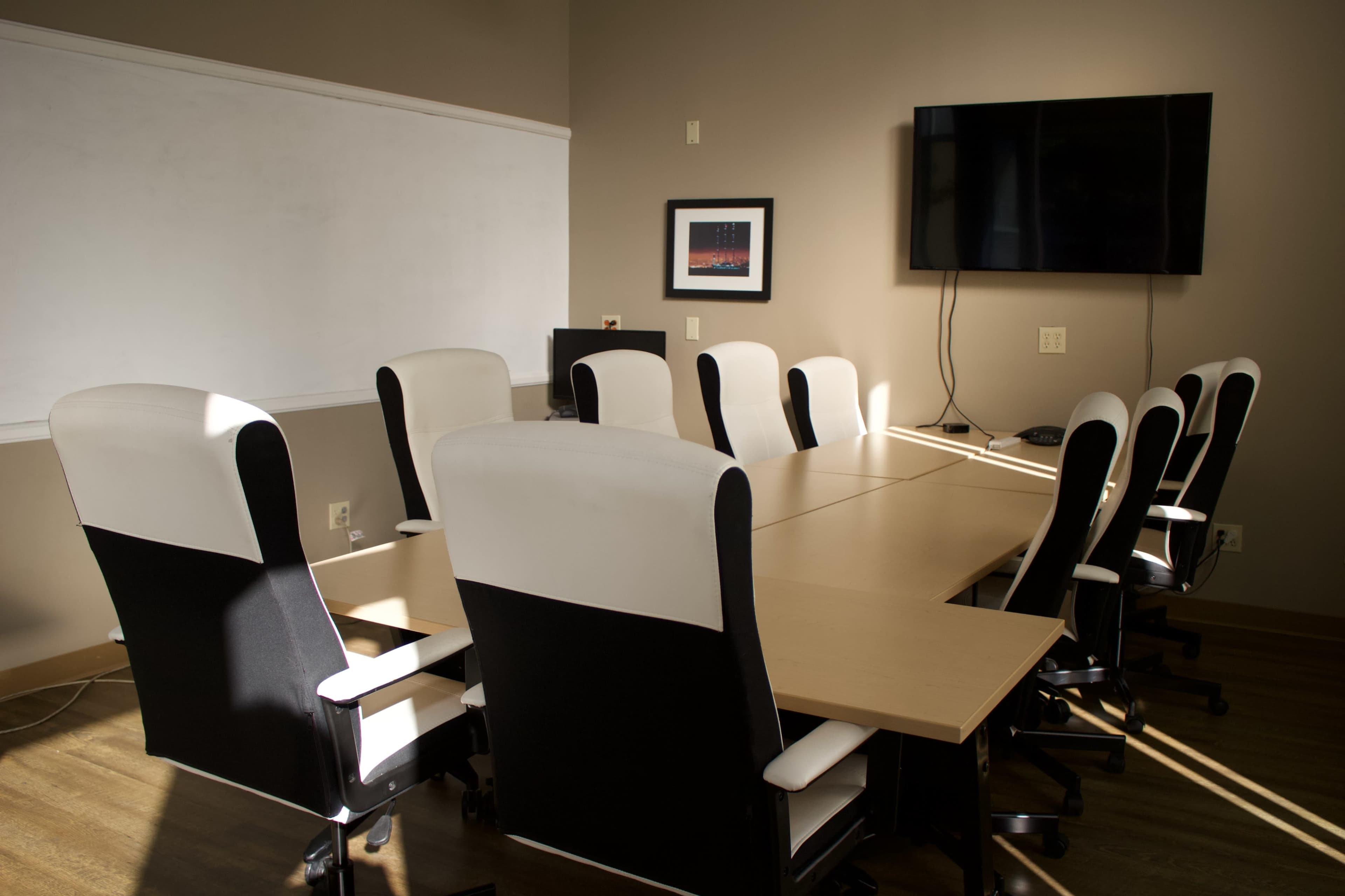 A modern conference room features a long table surrounded by ergonomic chairs, with a whiteboard and a wall-mounted television in the background.