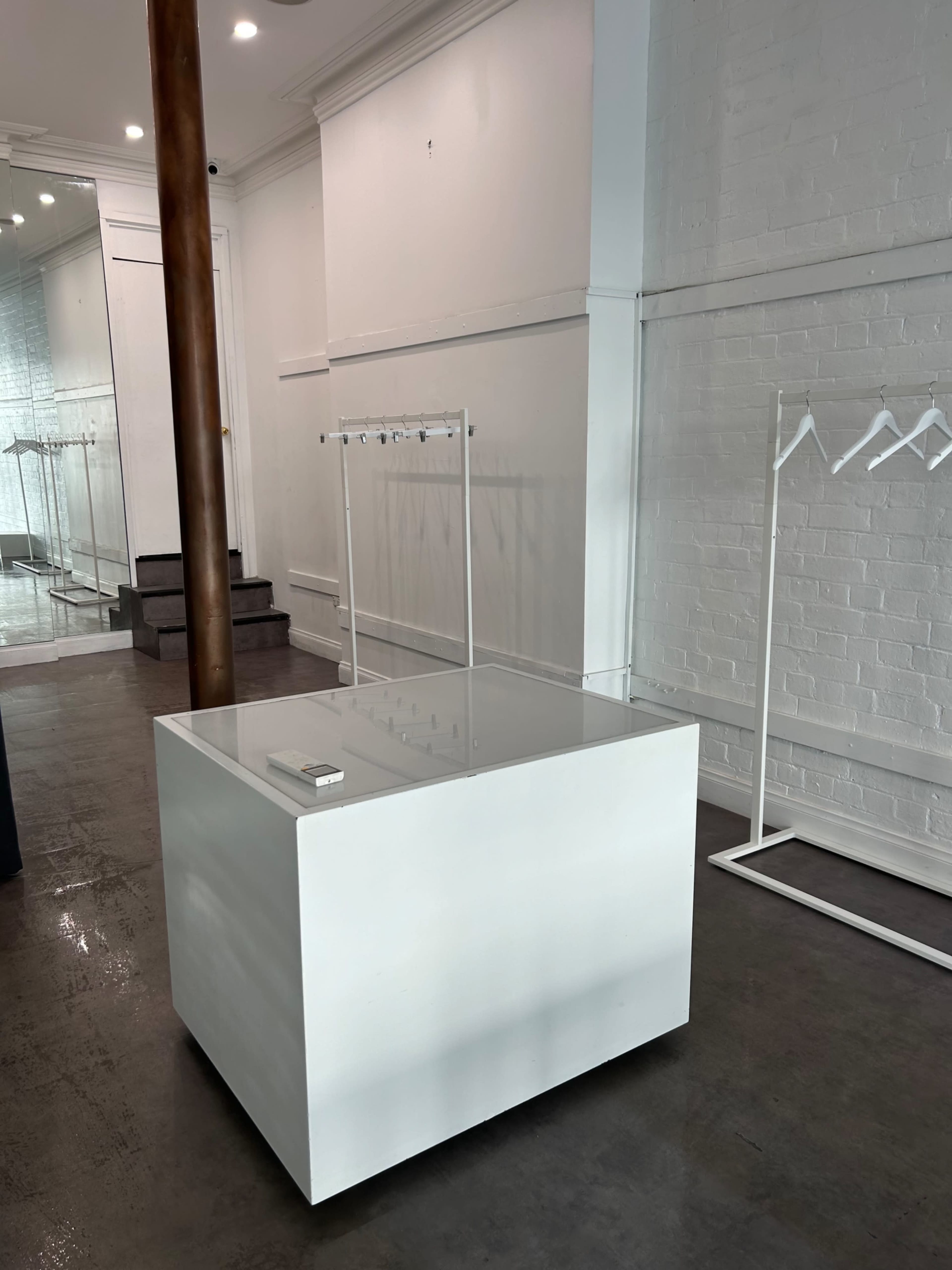 The image shows a minimalist retail space with a white display table and clothing racks against a light-colored wall.