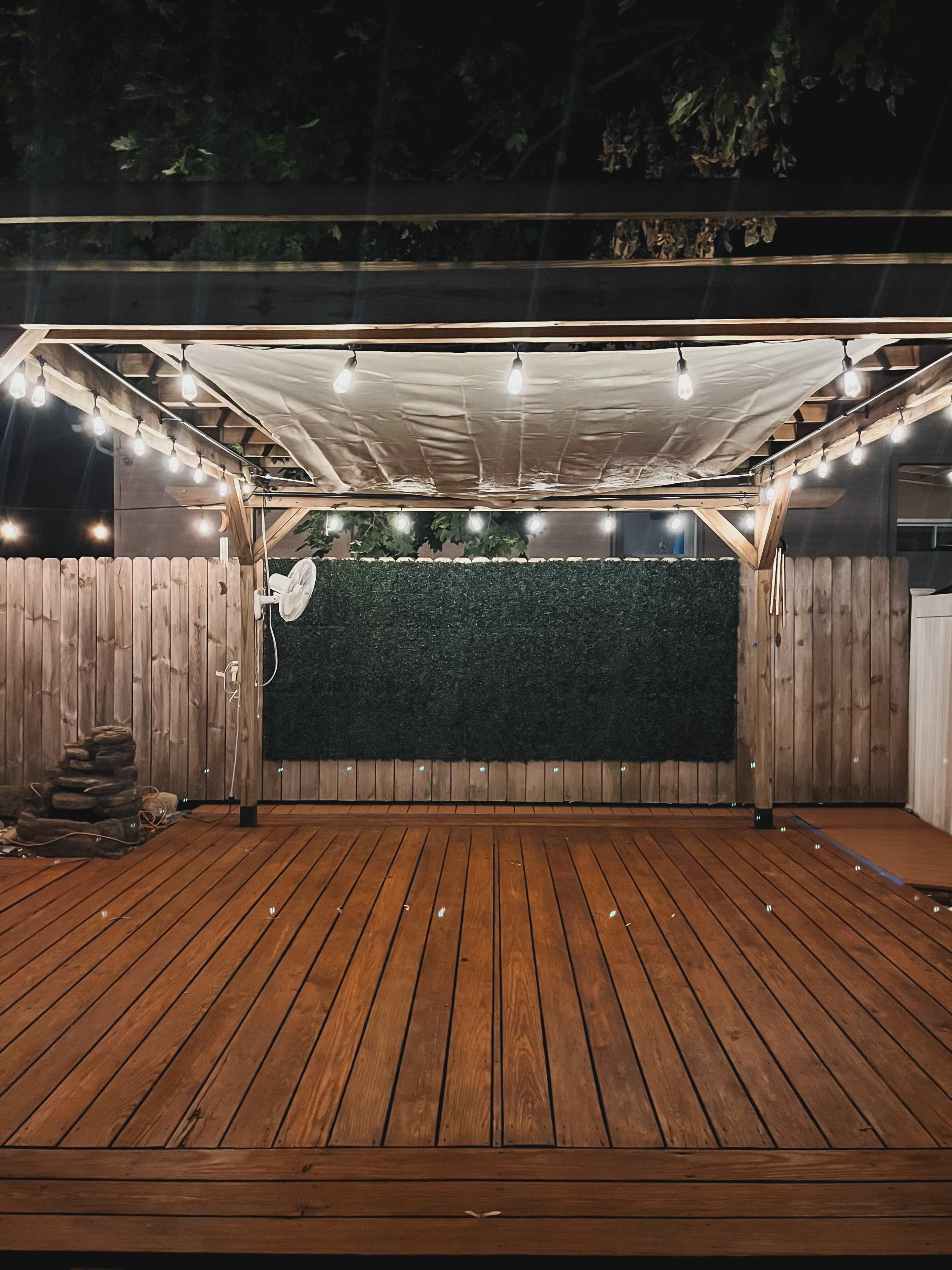 The image shows a wooden deck under a shaded pergola with string lights and a backdrop of artificial greenery.