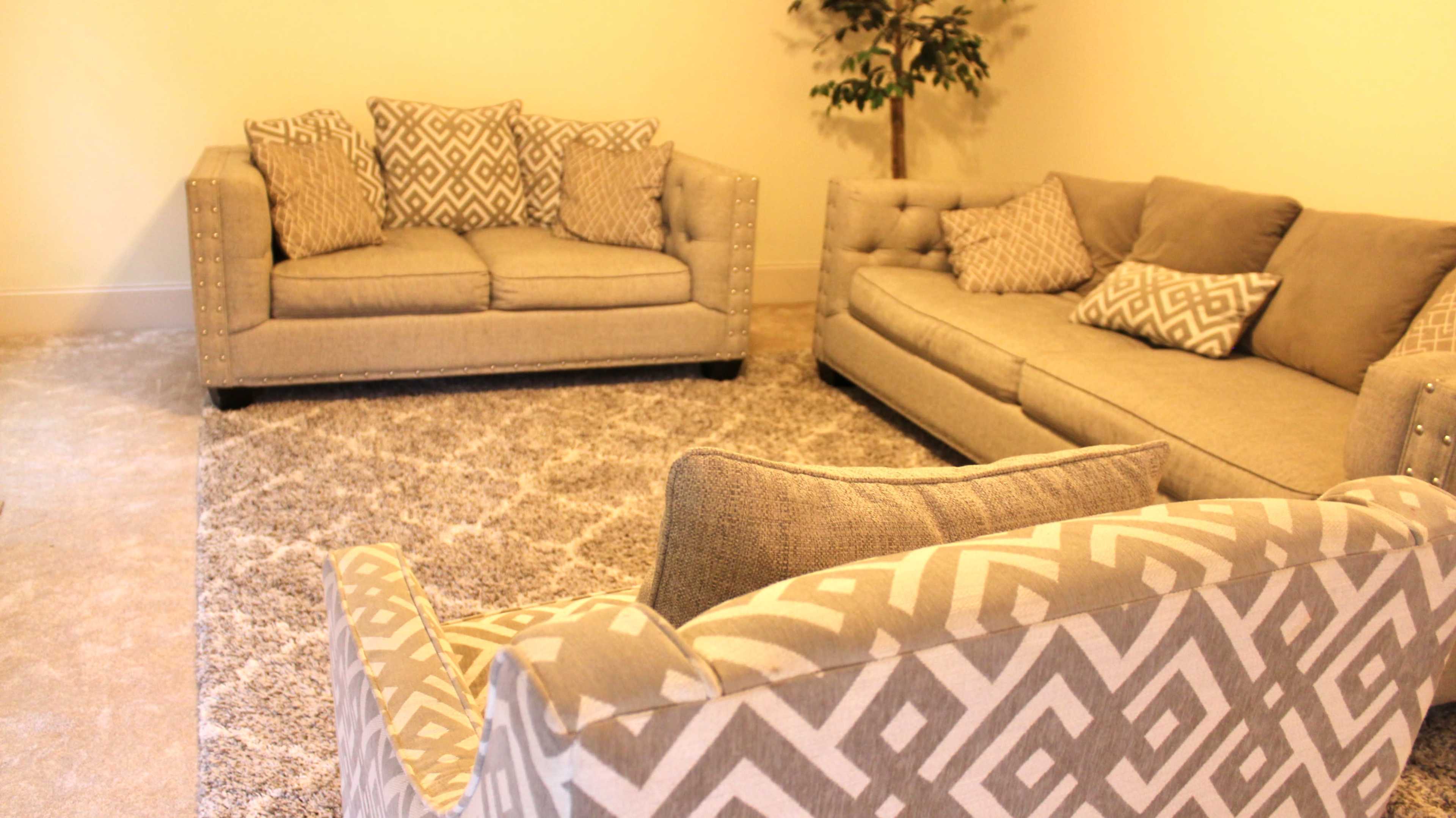 The image shows a living room with two couches and a patterned rug on a carpeted floor, featuring decorative pillows on each couch.