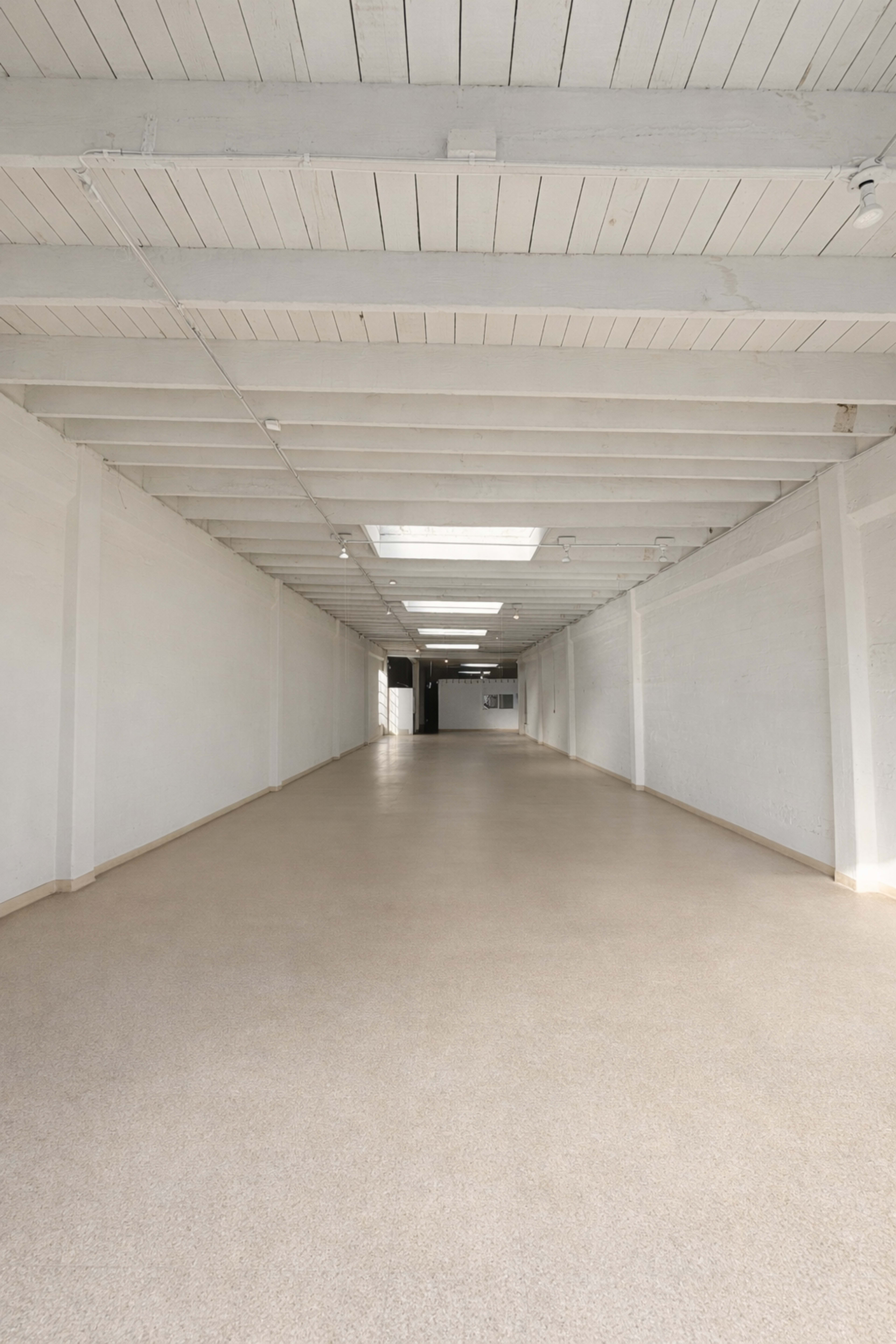 The image shows a spacious, empty room with a smooth beige floor, white walls, and exposed wooden beams on the ceiling.
