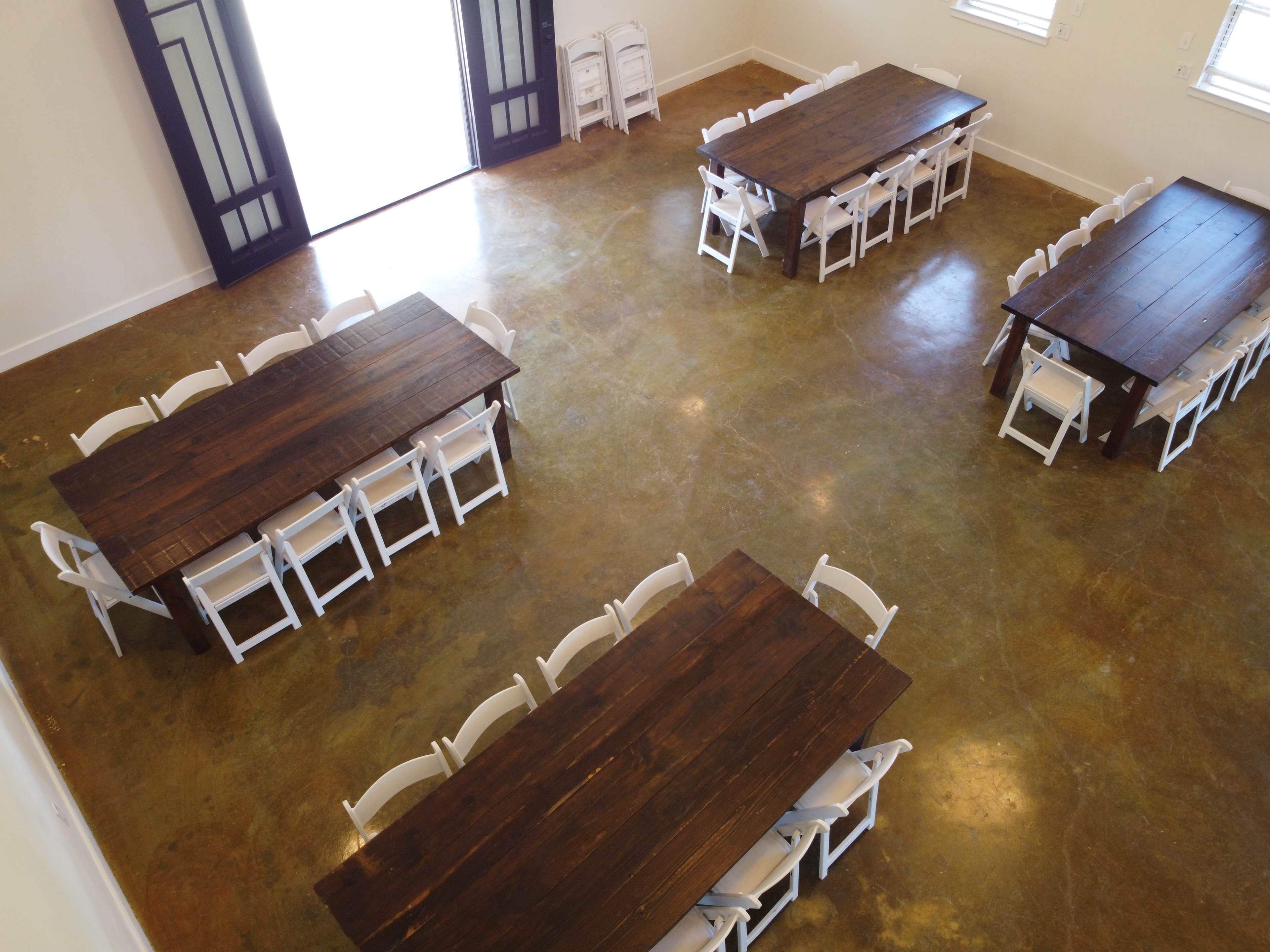 The image shows a well-lit room with four wooden tables arranged in a central layout, surrounded by white folding chairs.