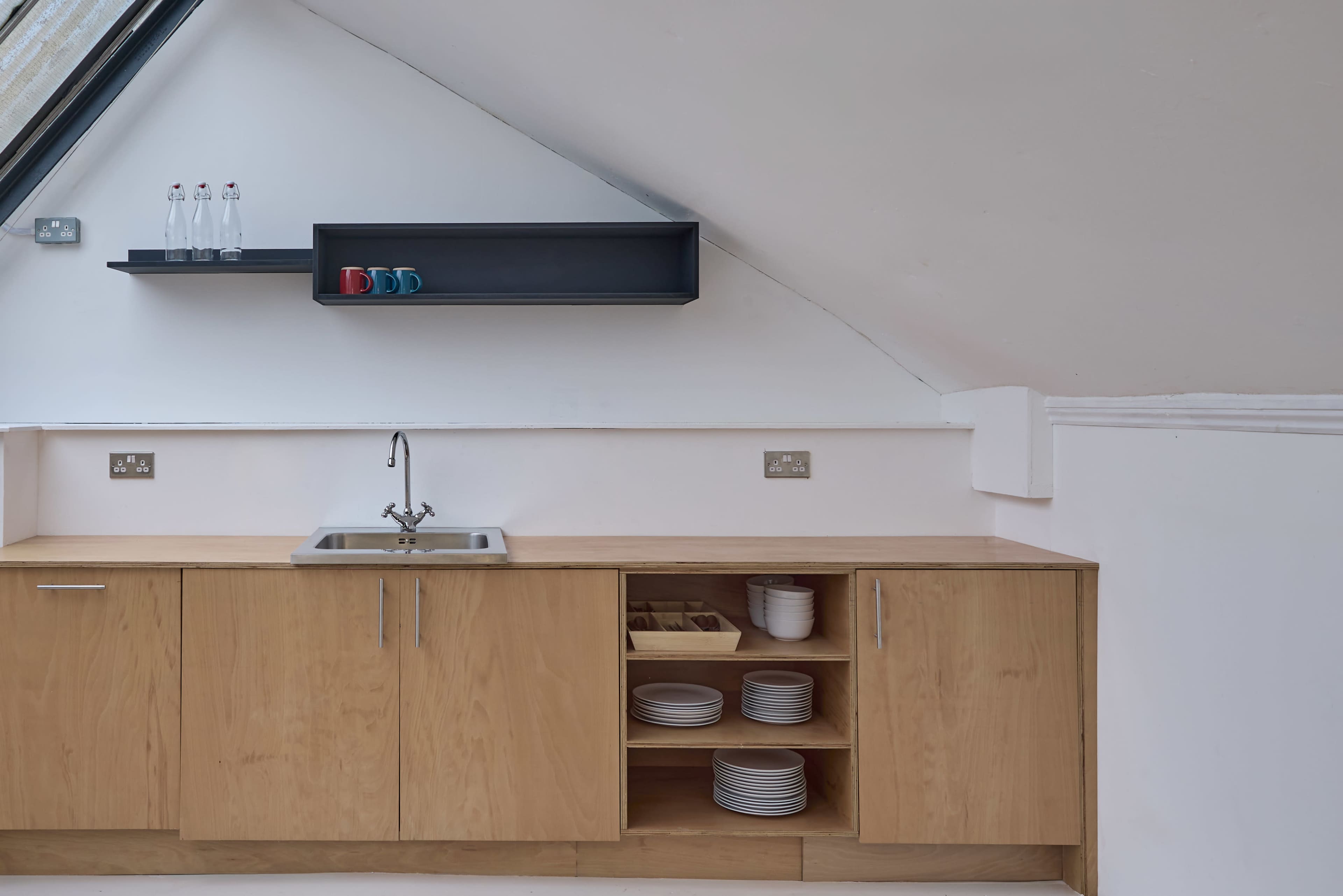 The image shows a modern kitchen area with wooden cabinetry, a stainless steel sink, and a shelf displaying bottles and colorful mugs.