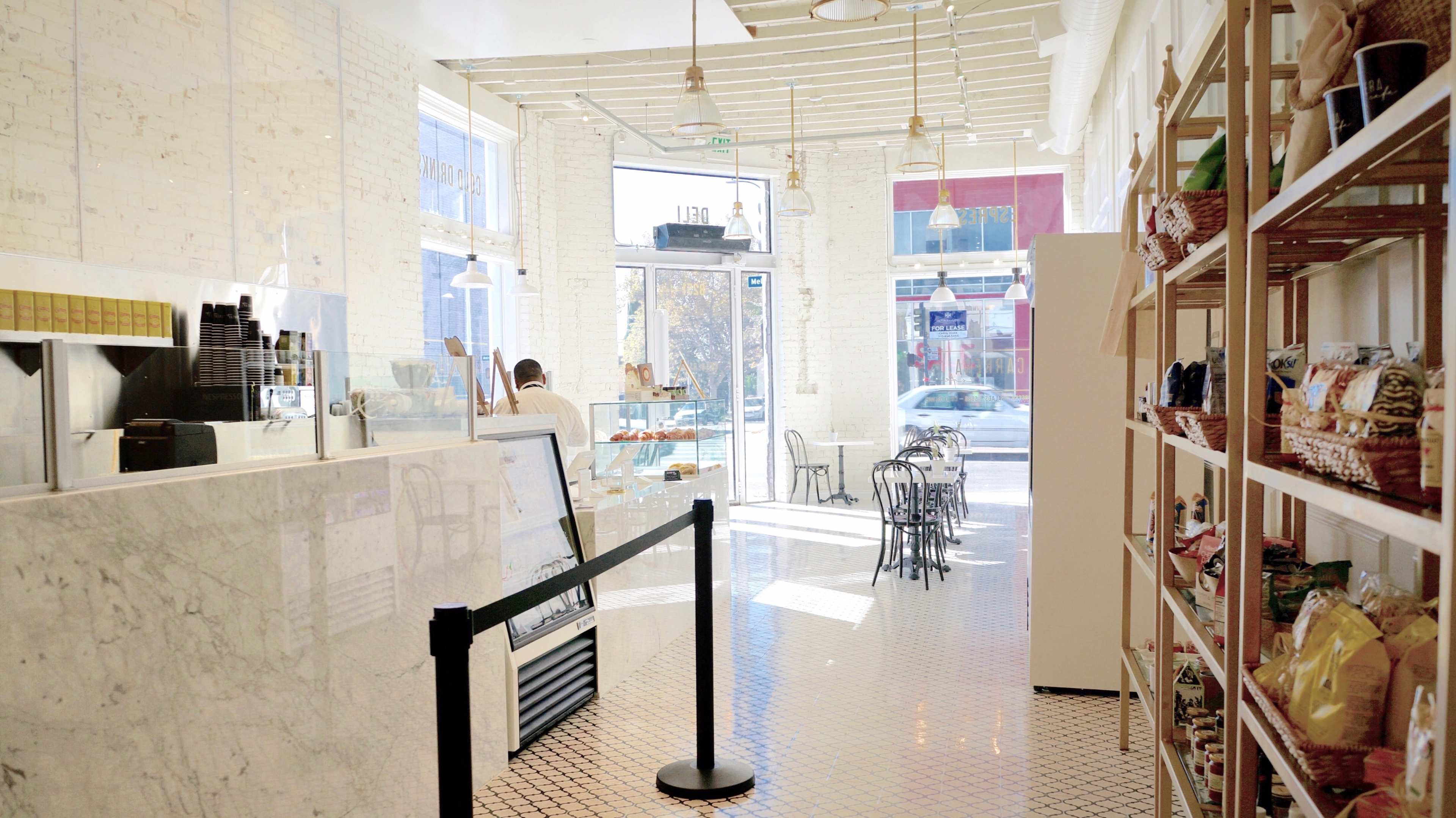 A bright café interior featuring a marble counter, black chairs, a display case with baked goods, and shelves filled with various products.