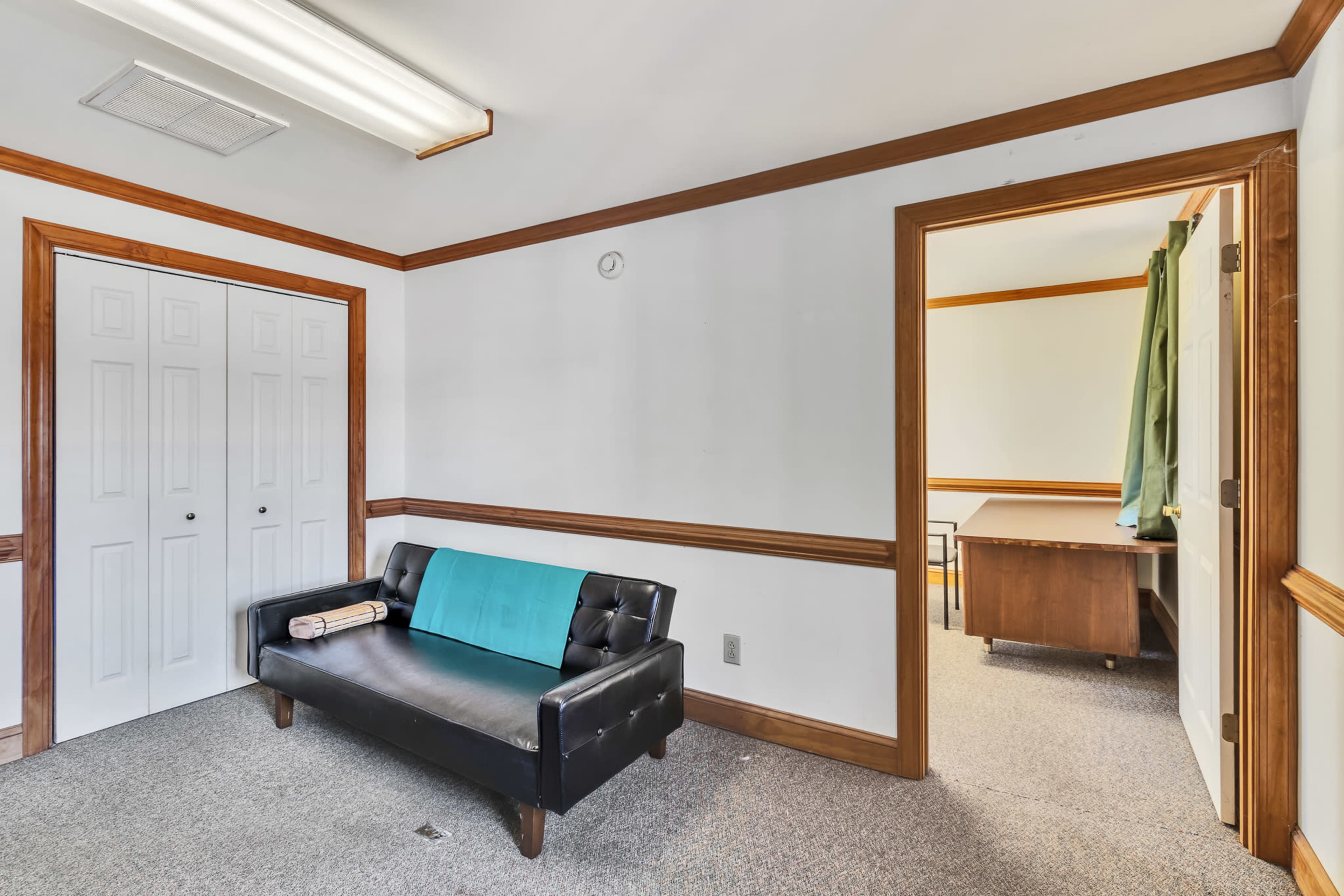 The image shows a small room with a black couch, a rolled-up item on the cushion, and a door leading to another room with a desk visible beyond.