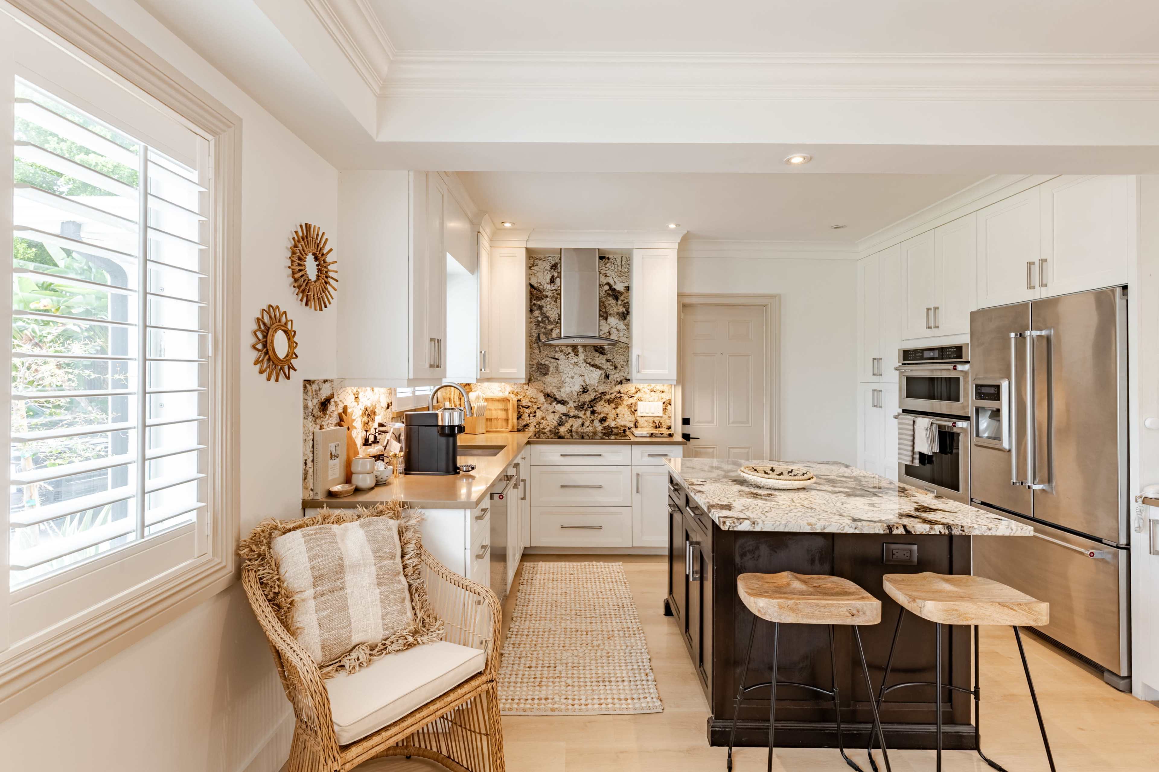 The image shows a modern kitchen featuring white cabinetry, a large island with dark wood accents, and a marble countertop, along with a cozy seating area with a woven chair.