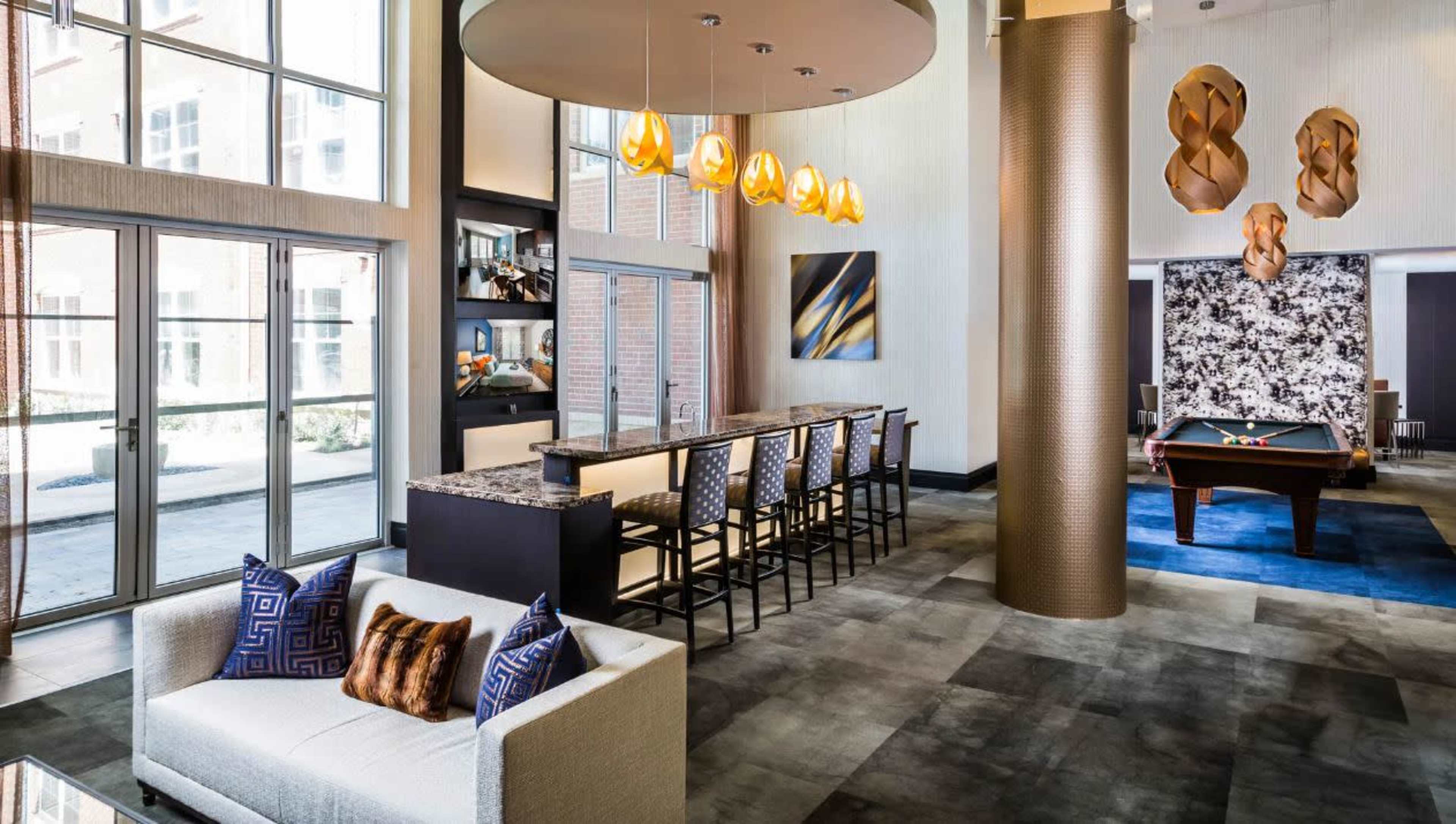 A modern lounge area featuring a bar with high stools, a pool table, and decorative lighting above.