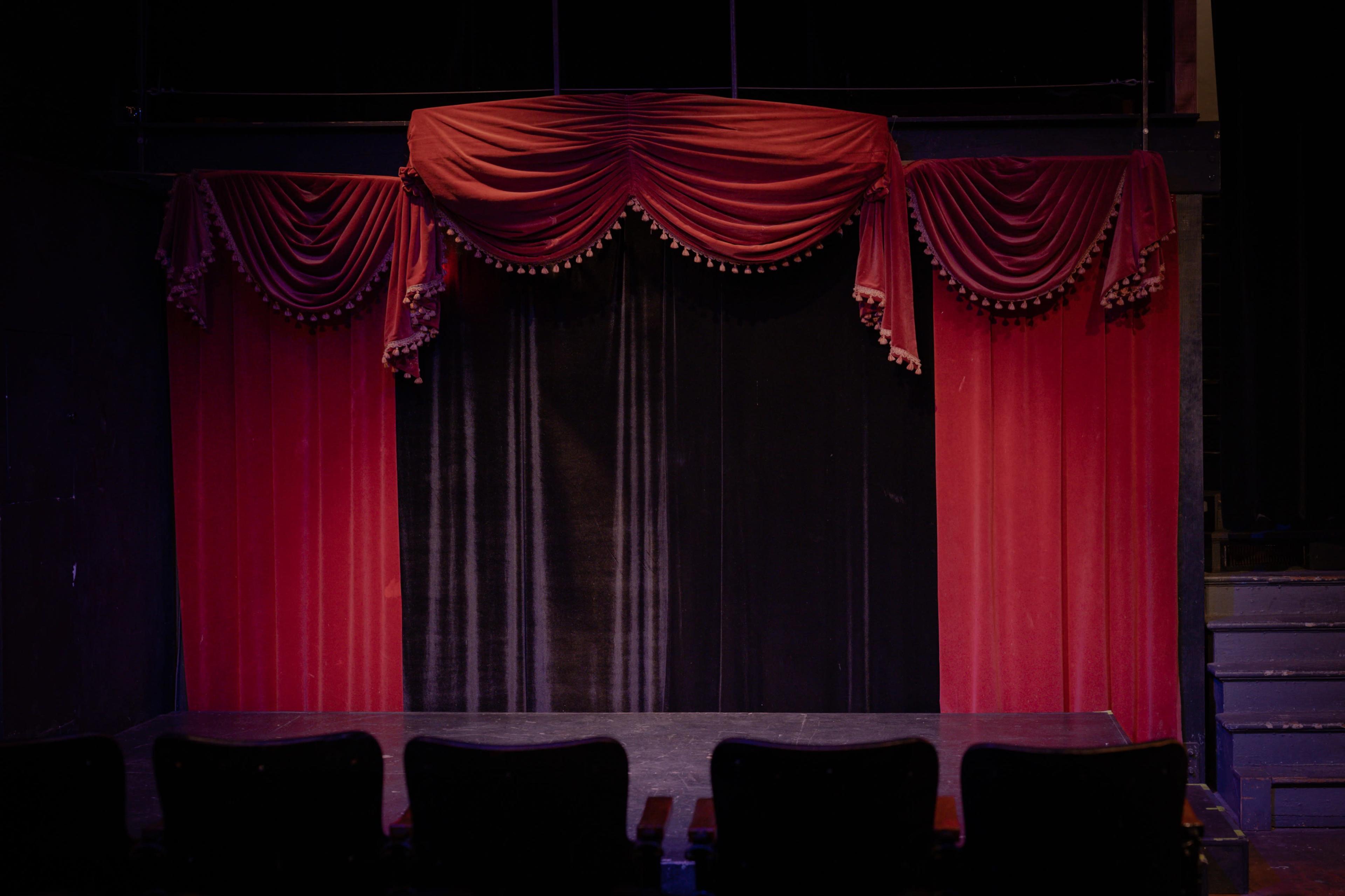 A theater stage draped with red curtains, framed by ornate valances, and with empty black seats in front.