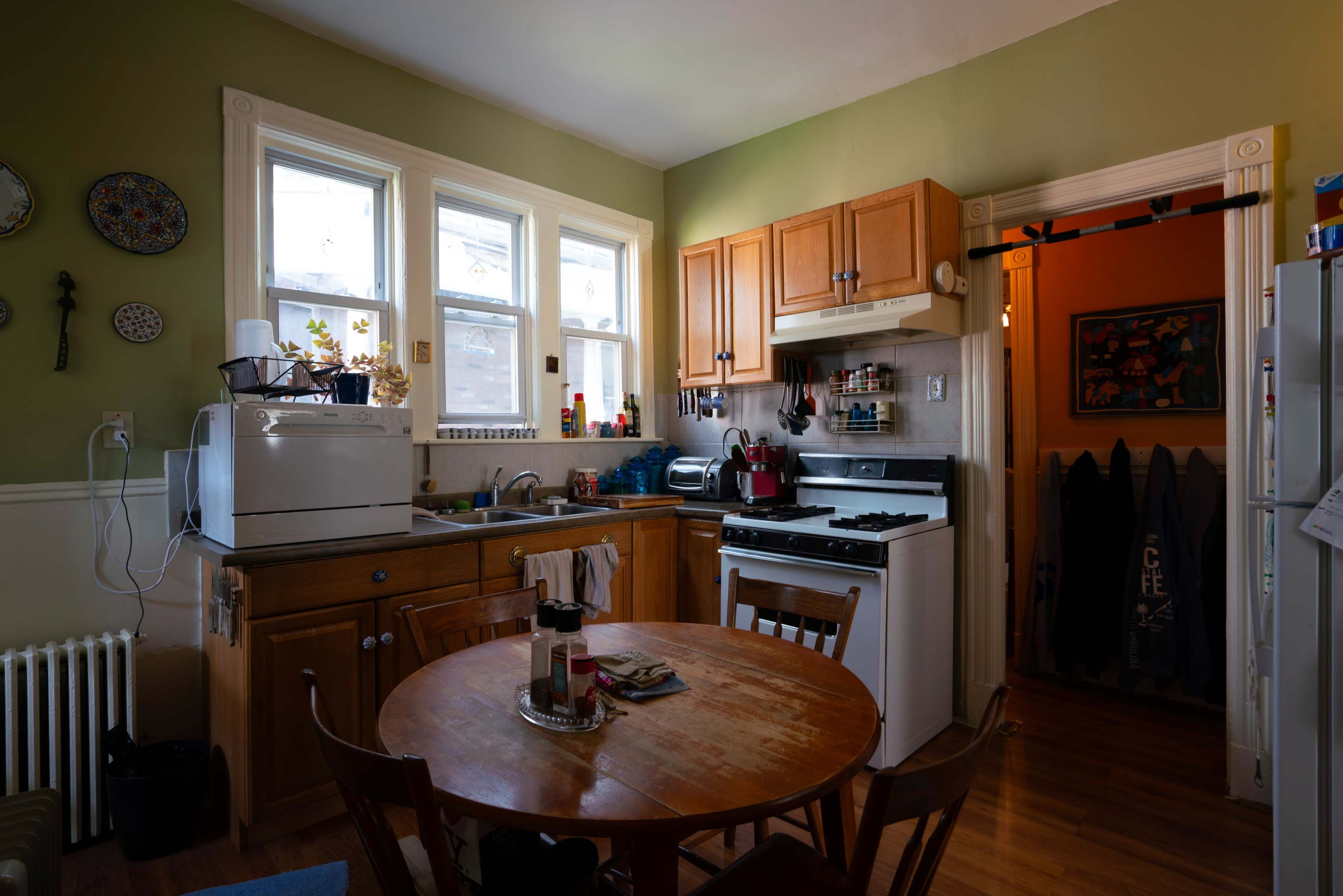 The image shows a small kitchen with wooden cabinetry, a dining table, and various kitchen appliances.