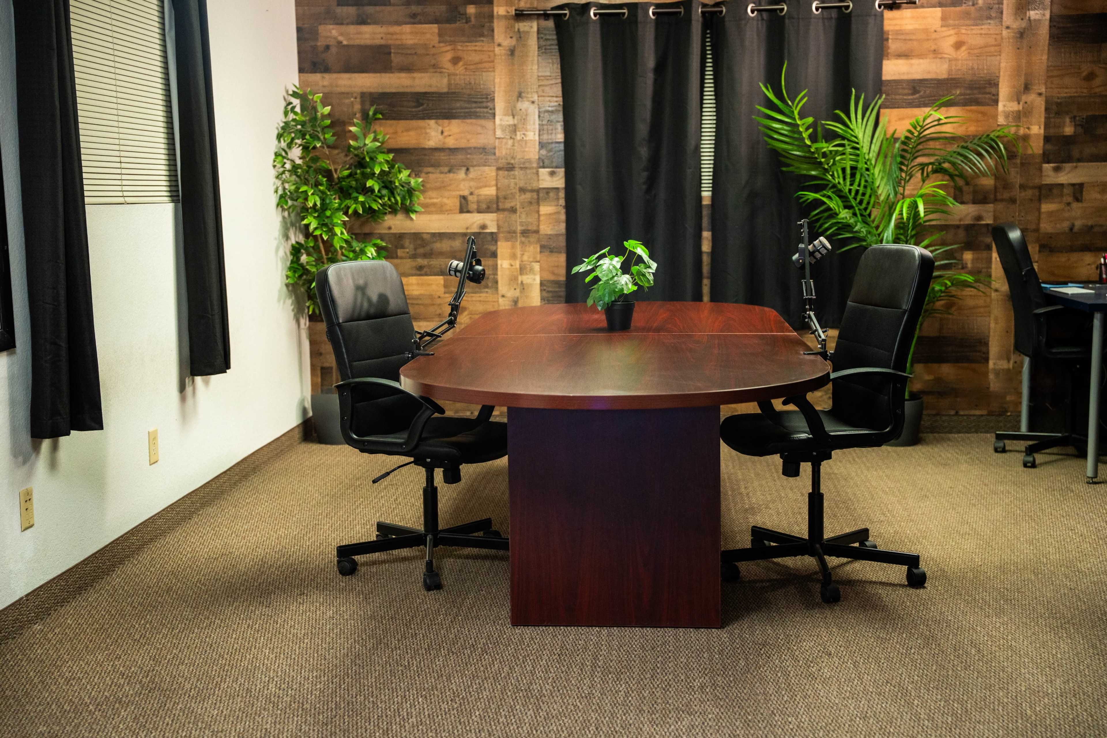 The image shows a conference room with a round wooden table, two black chairs, and greenery against a backdrop of wooden paneling and dark curtains.