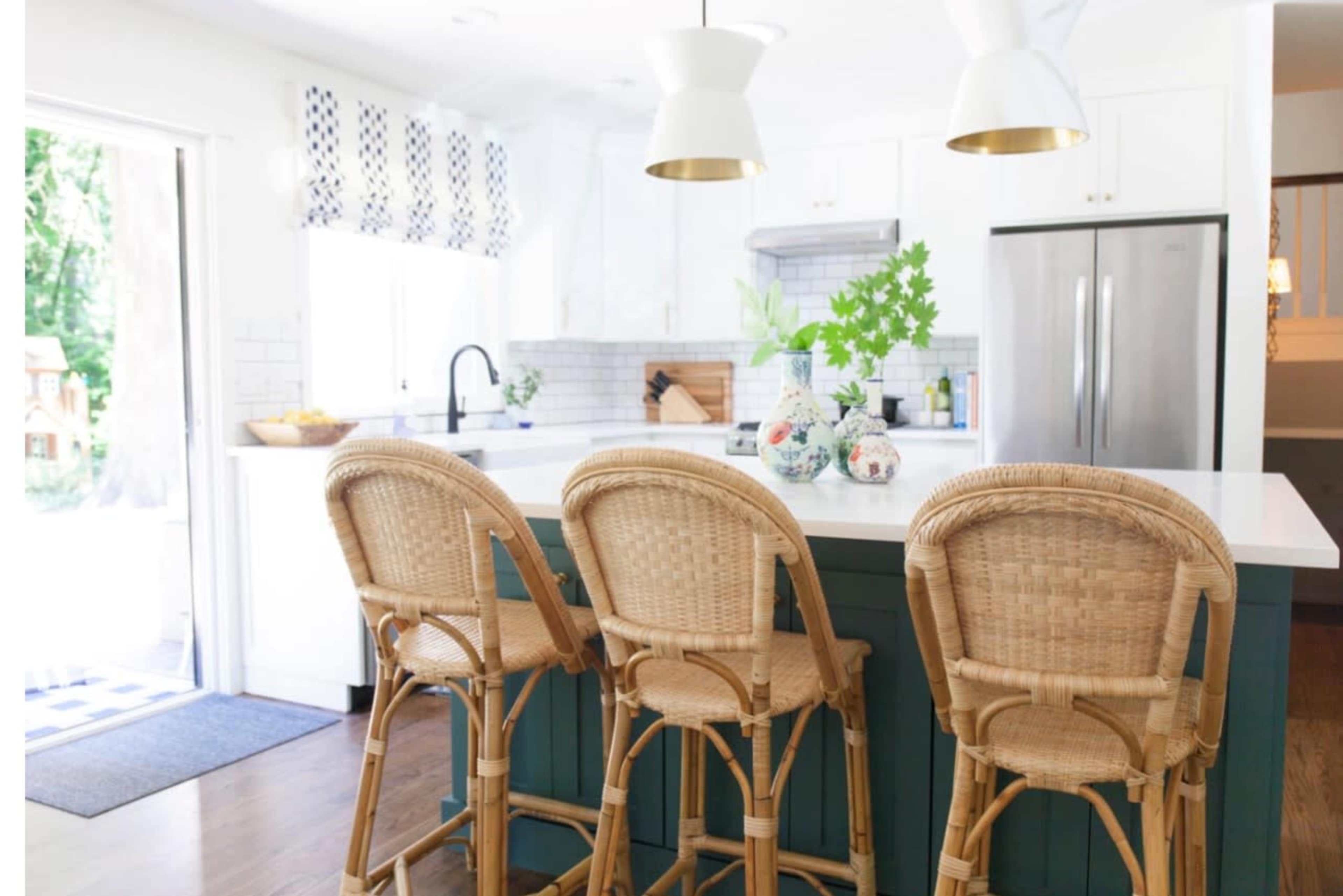 A modern kitchen features a large island with rattan bar stools, white cabinetry, and a view of an outdoor area through an open door.
