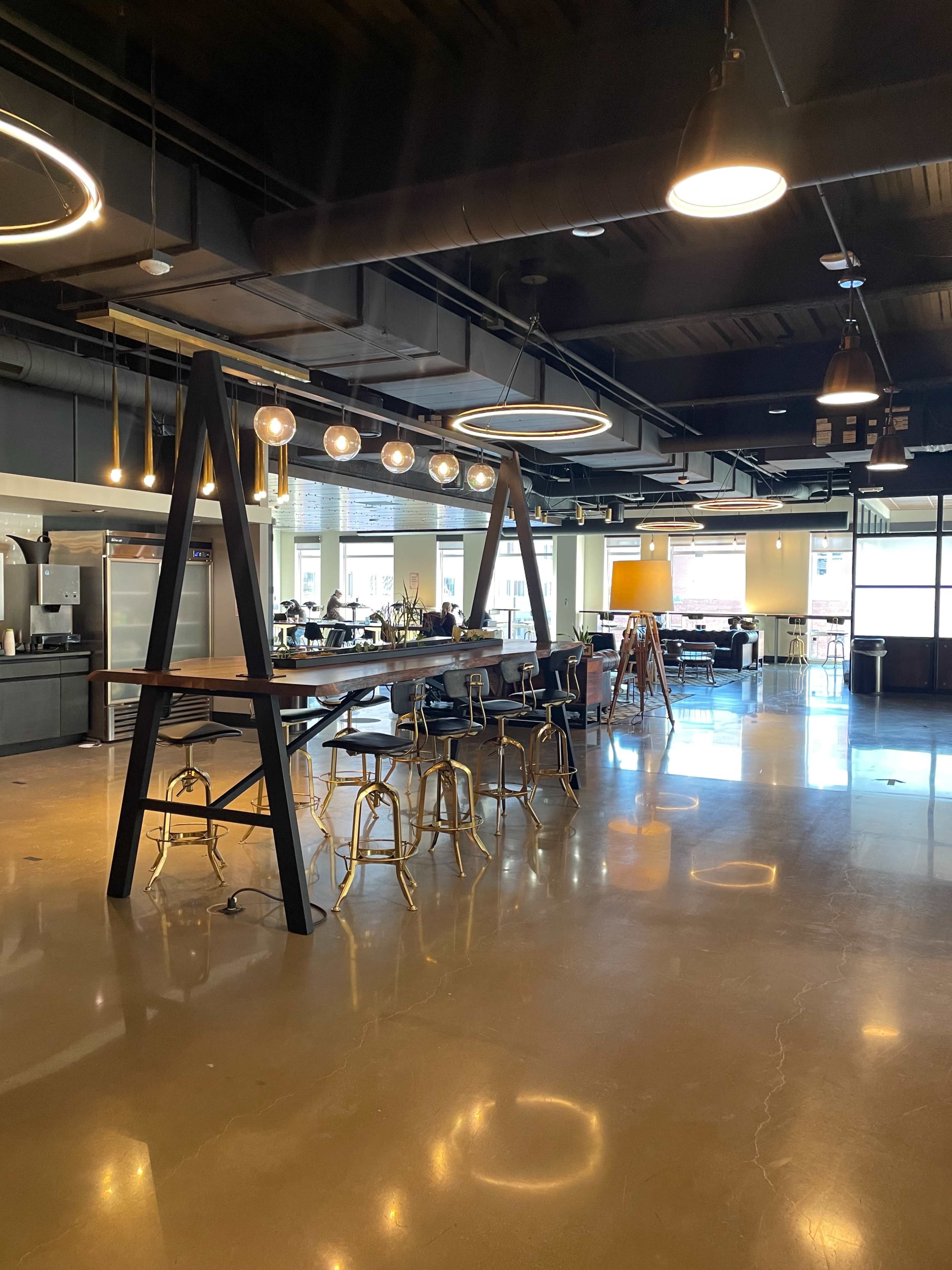 A modern office space with high ceilings, a large wooden table supported by black A-frame legs, and pendant lighting.