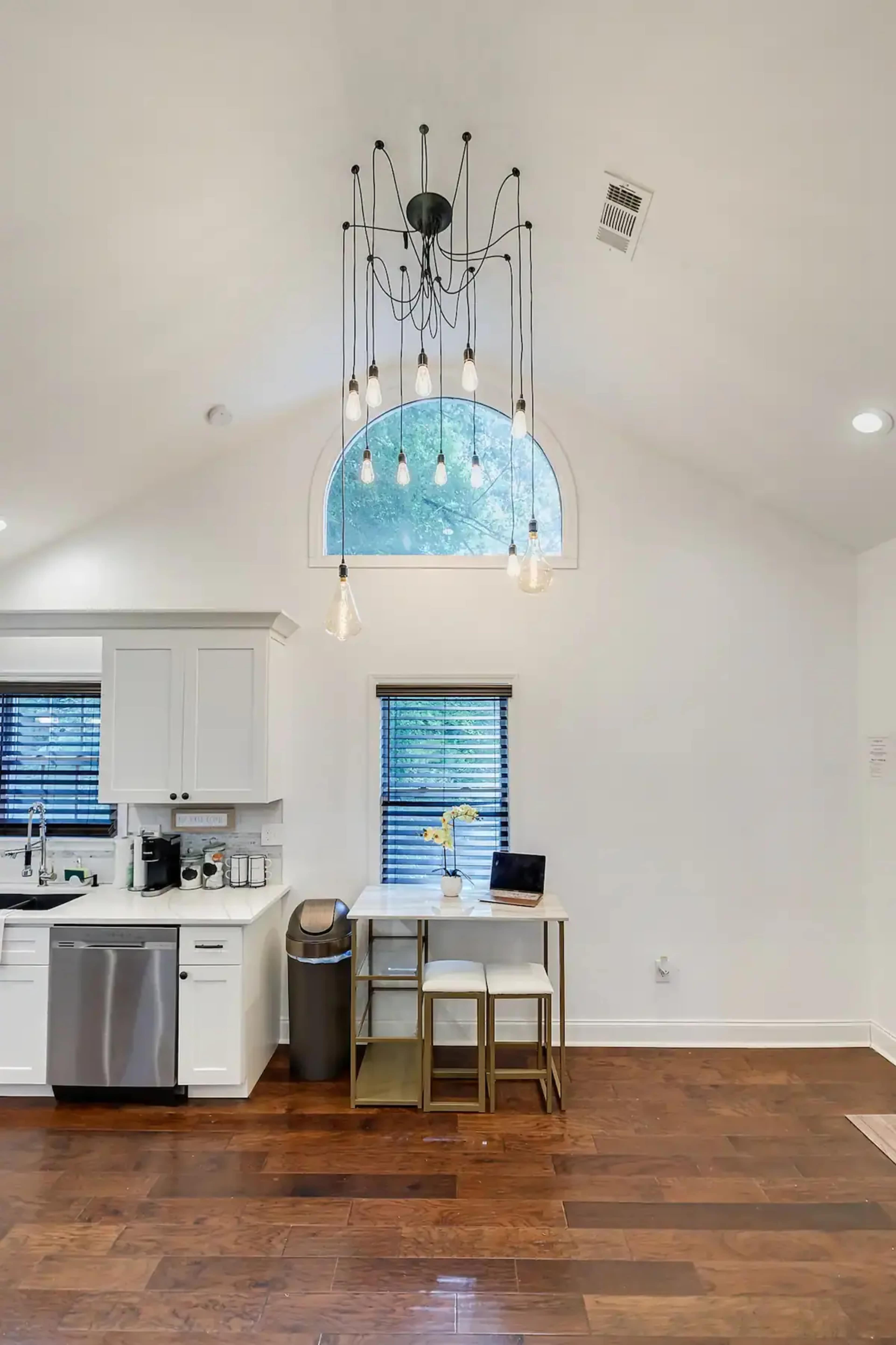 A modern kitchen area with white cabinetry, a small dining table with two stools, and a large arched window above, surrounded by wooden flooring.